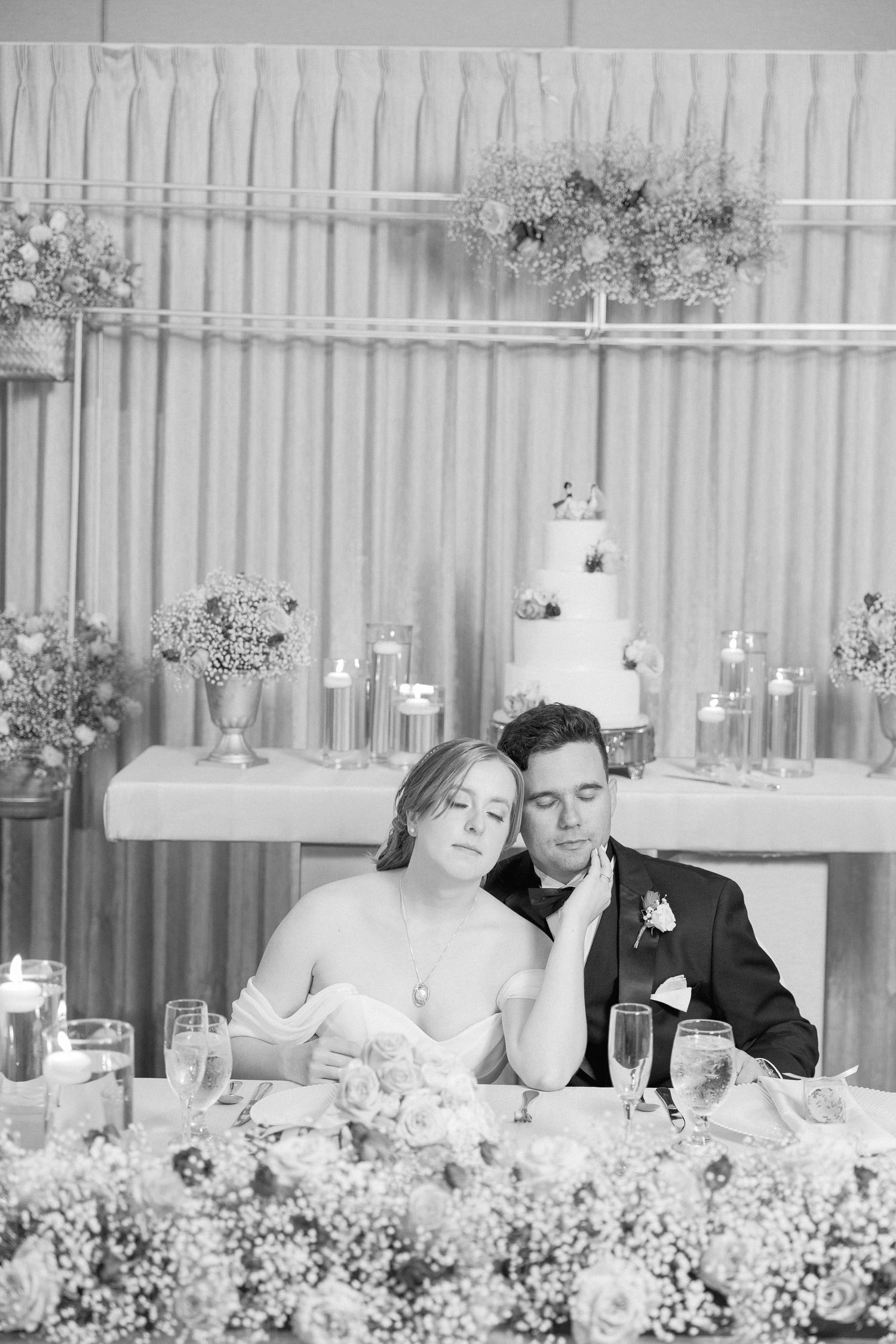 A romantic wedding reception scene with a tiered white cake and floral decorations in the background.