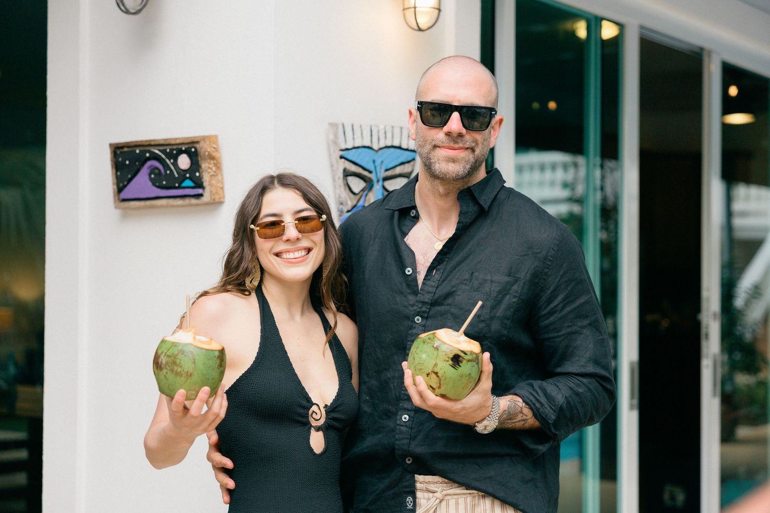 A couple in sunglasses holding fresh coconuts outside a storefront.