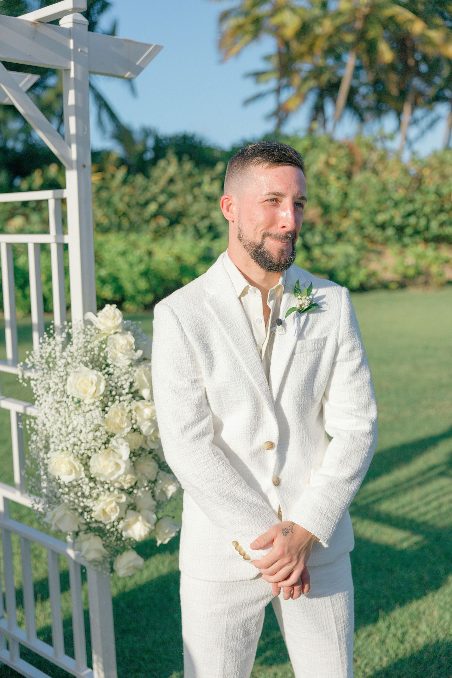 White floral arrangements adorn a wedding arch on a sunny lawn with palm trees.