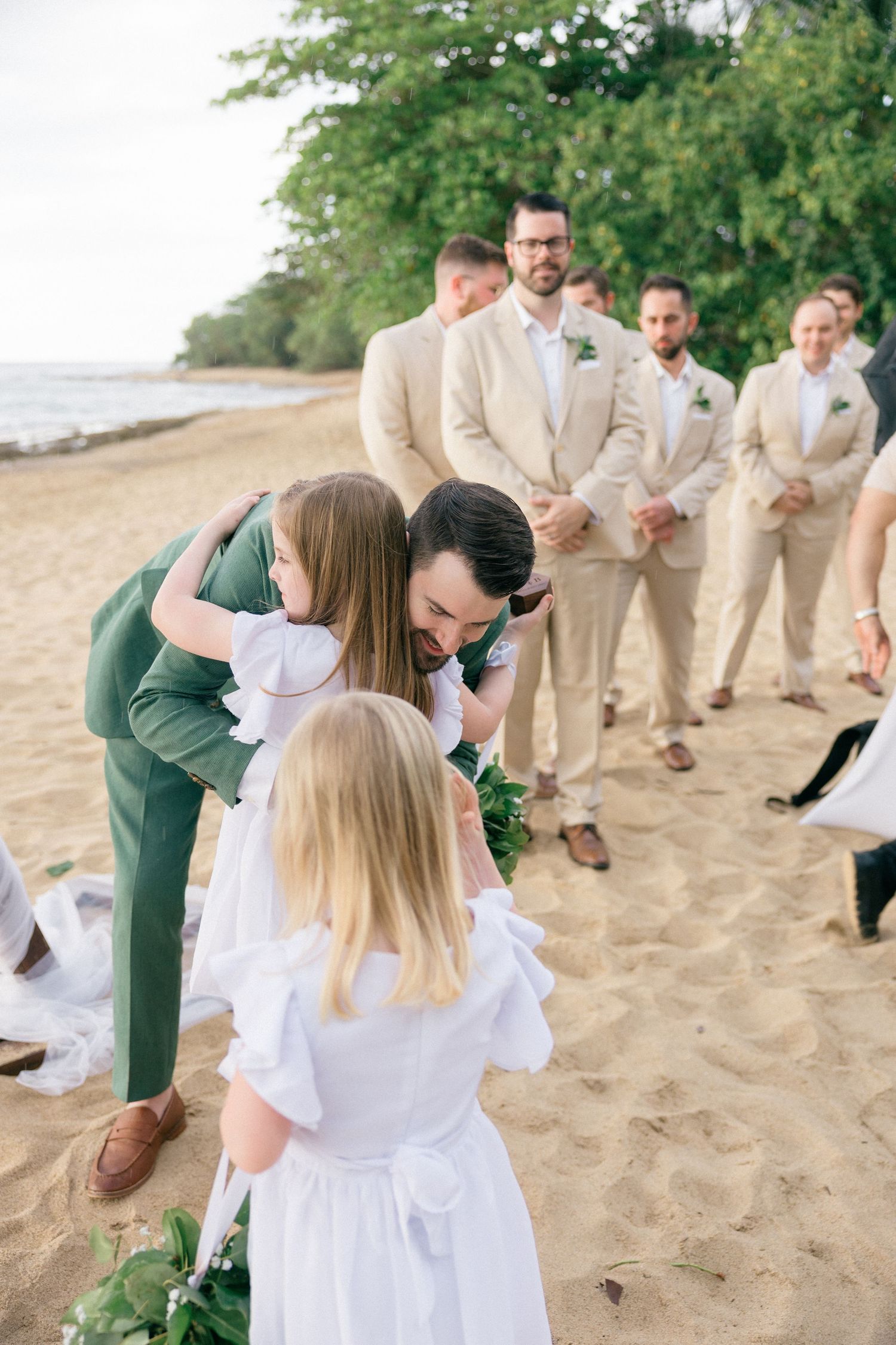 Wedding party members interact with flower girls on a sandy beach during ceremony.