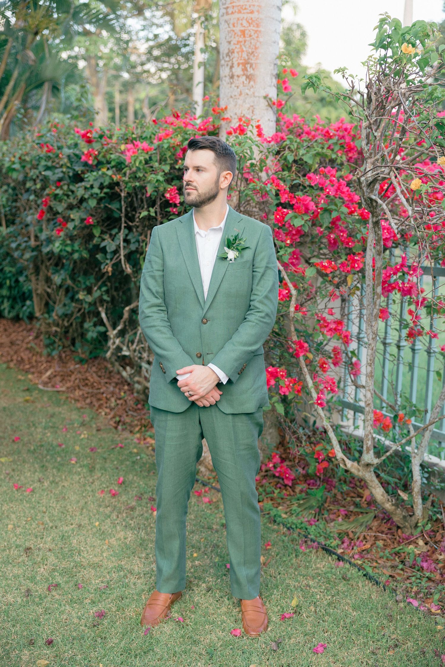 Groom in sage green suit poses against backdrop of red flowering bushes in tropical garden.