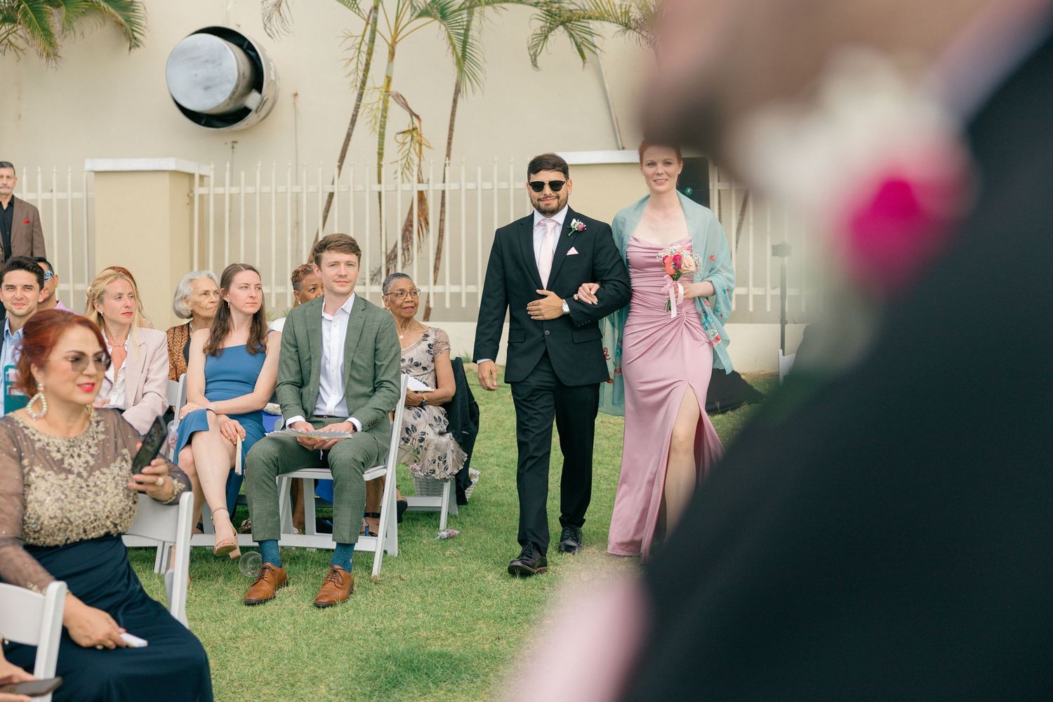 Wide shot of wedding ceremony seating arrangement with tropical landscaping and white chairs.