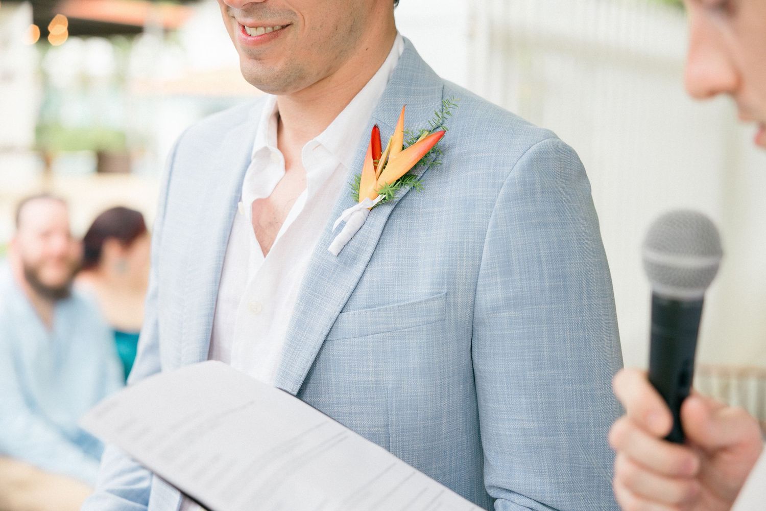 Groom in light blue suit jacket with tropical flower boutonniere reads from paper during outdoor wedding ceremony.