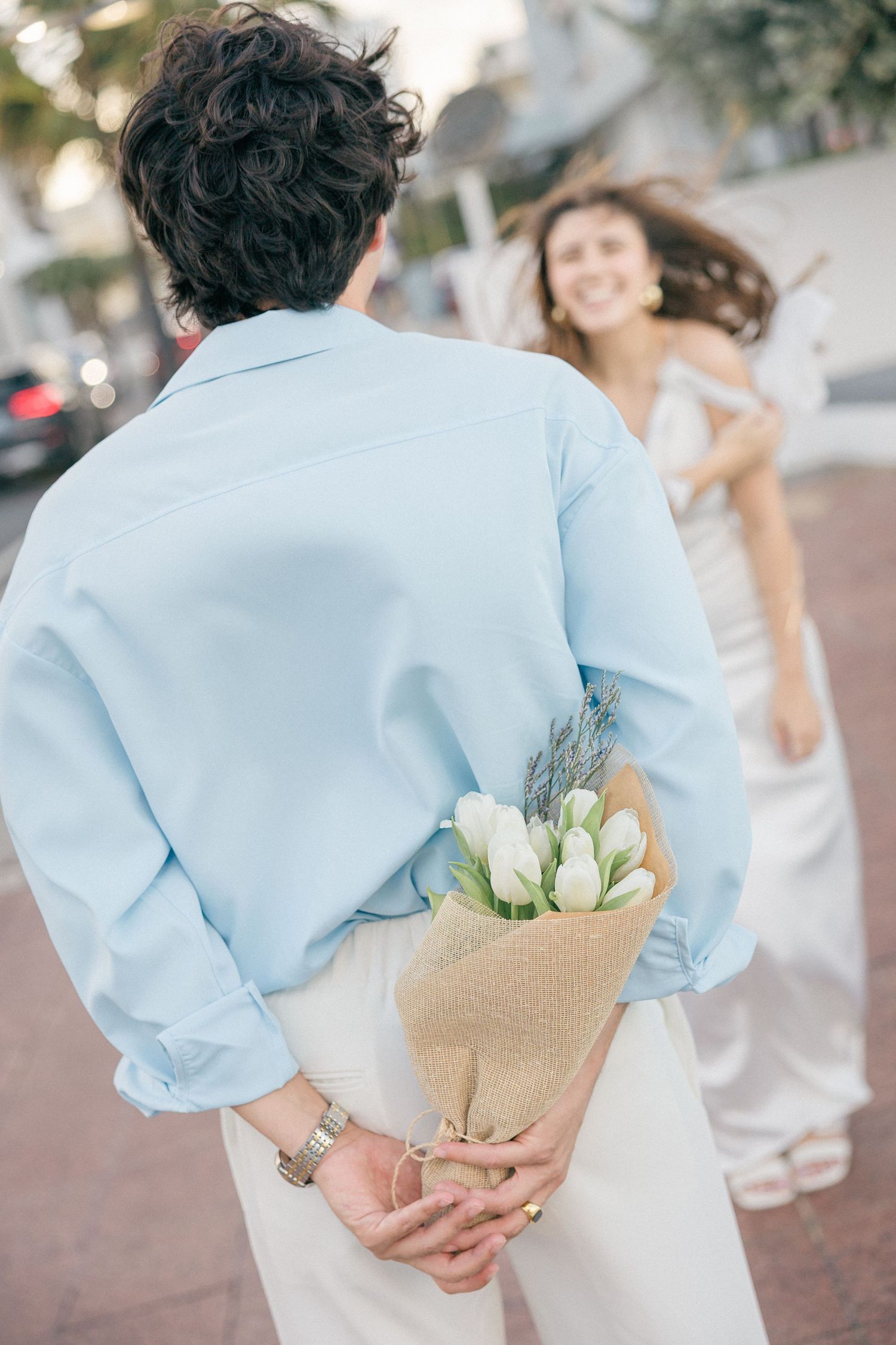 Person in blue shirt hiding bouquet of white tulips behind back as partner twirls in white dress.
