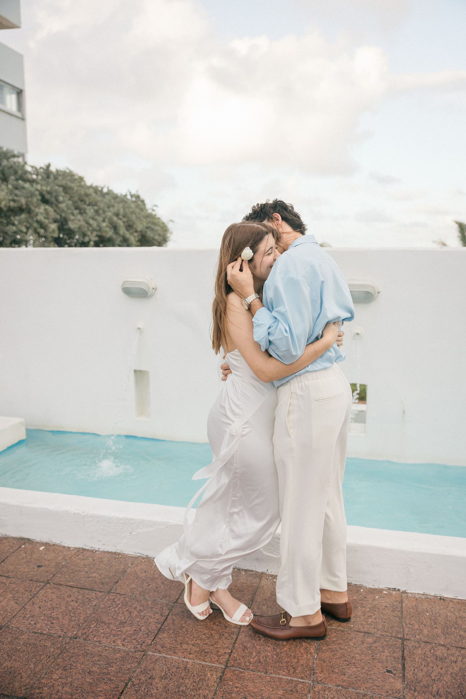 Couple in white and blue outfits embracing by a poolside.