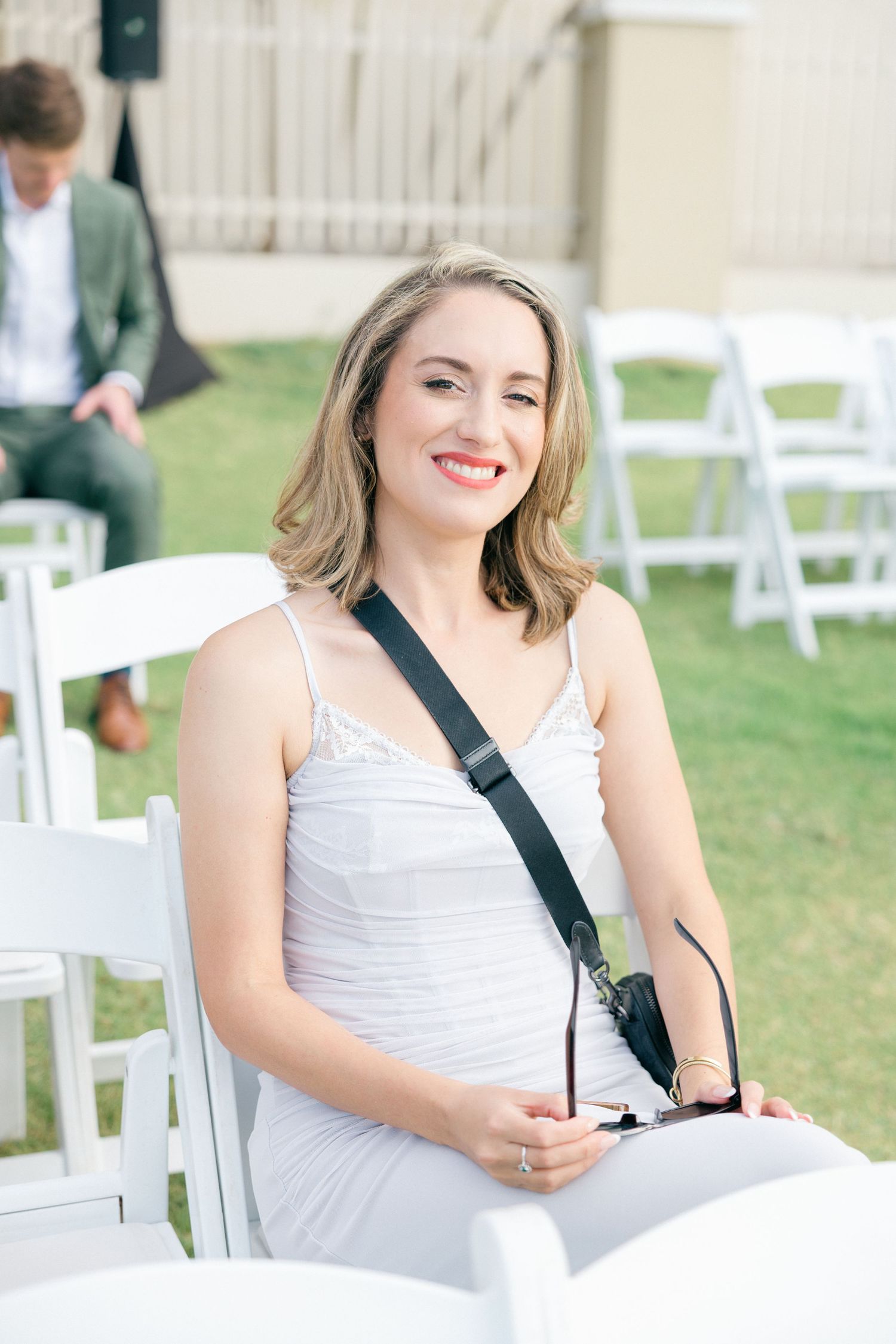 Professional photographer in white dress with camera strap sits in white folding chair outdoors.