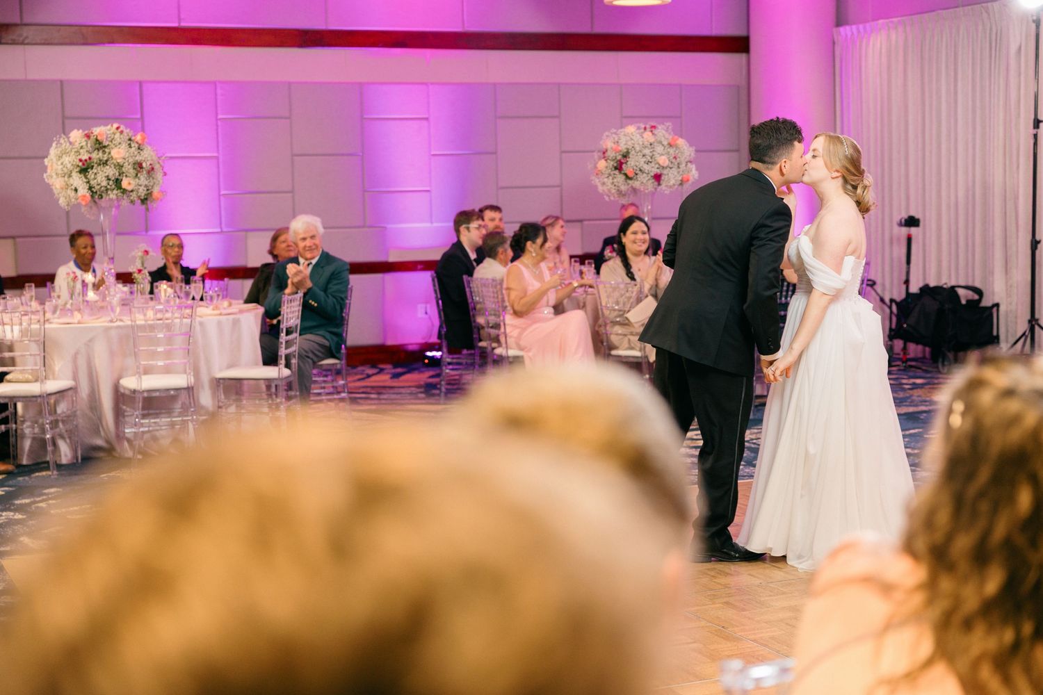 Wedding reception venue decorated with pink uplighting and white floral centerpieces as couple dances on the floor.