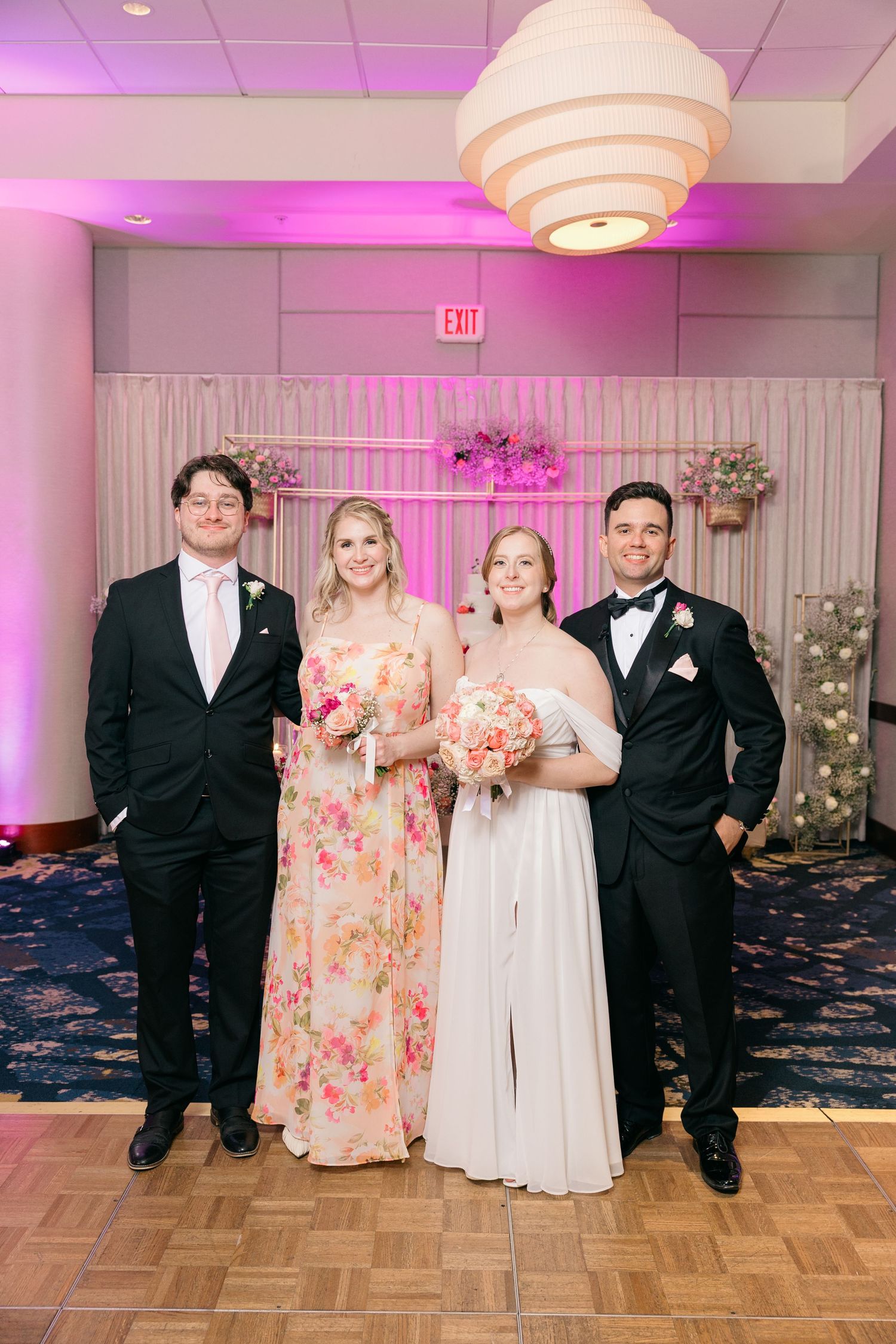 Wedding party members pose in formal attire against a pink-lit backdrop with floral arrangements.
