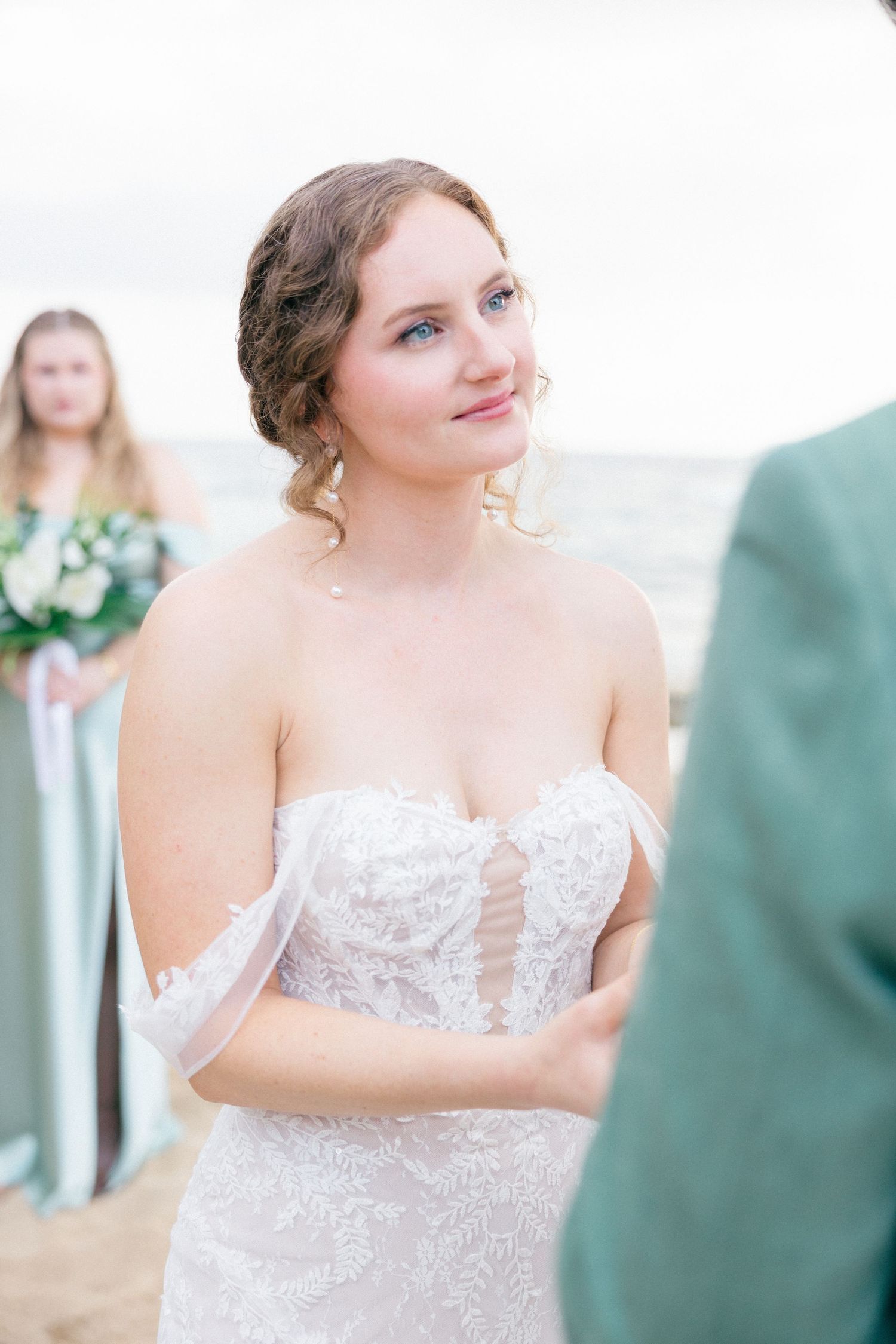 Bride in strapless lace gown smiles during seaside wedding ceremony.
