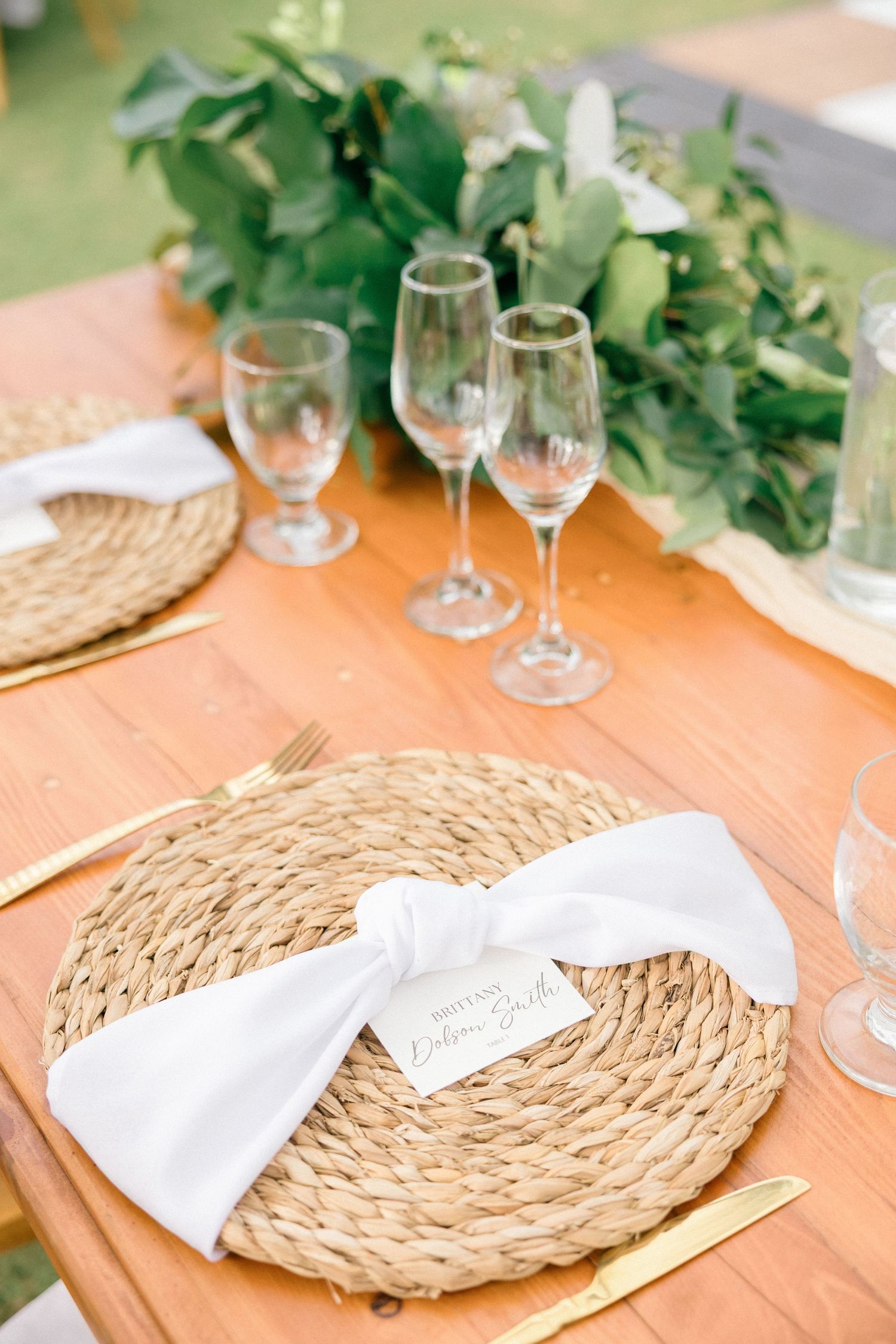 Rustic table setting with woven chargers, white napkins and greenery accents.