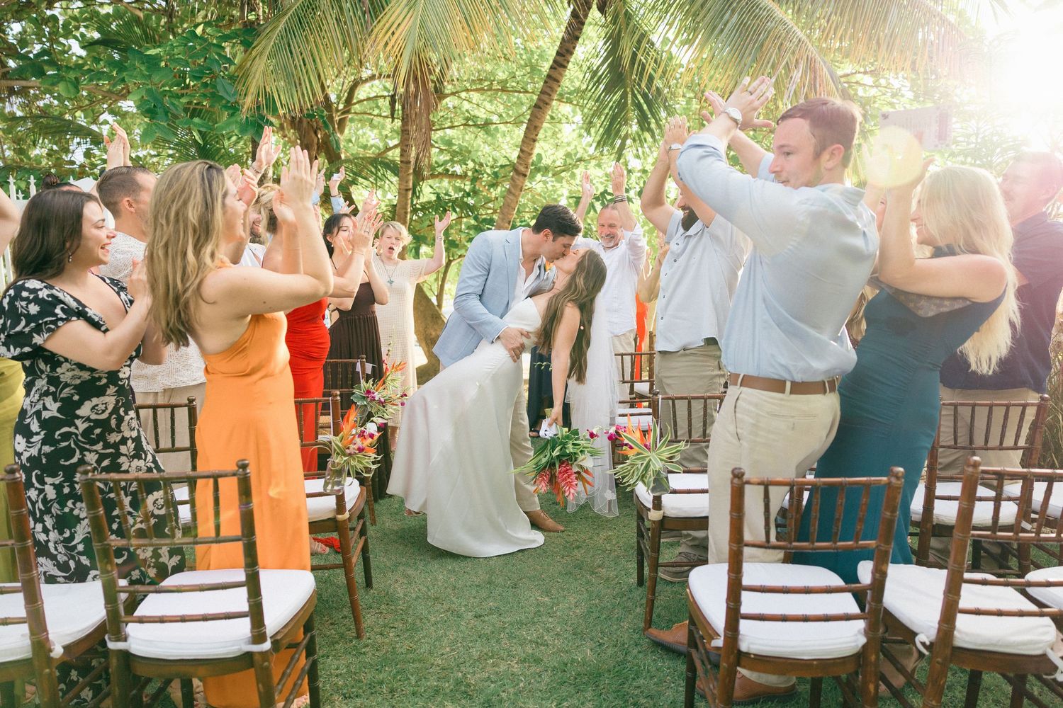 Outdoor wedding reception with guests seated in wooden chairs under natural sunlight.
