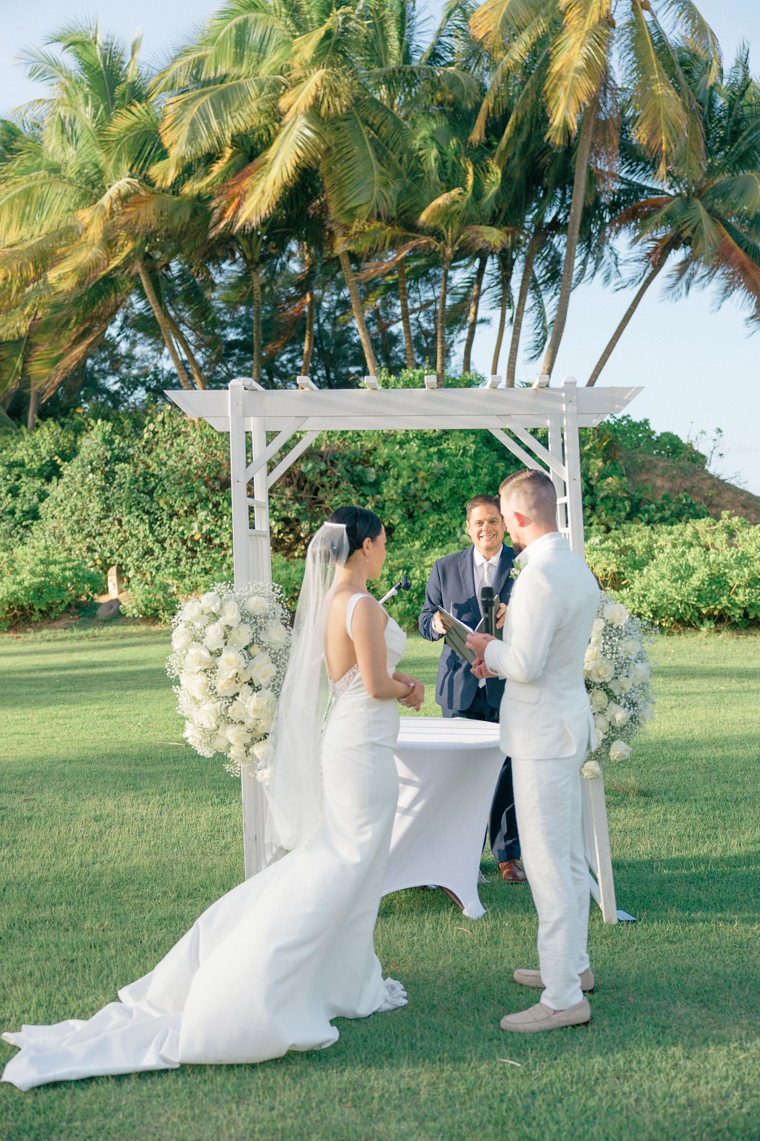 Tropical beach wedding ceremony with white arbor and palm trees swaying in the breeze.