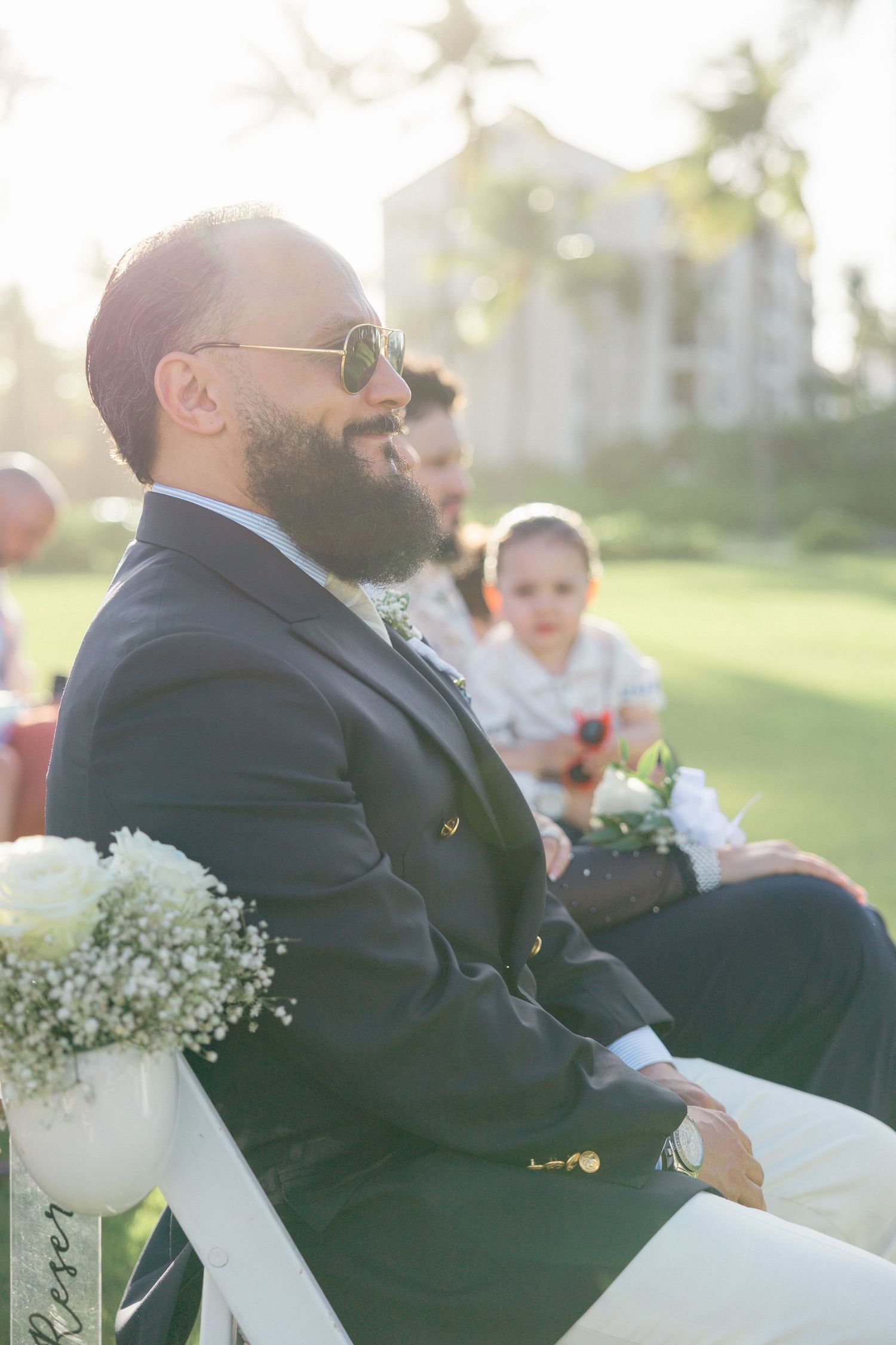 Wedding guests in formal attire seated in white chairs during outdoor ceremony.