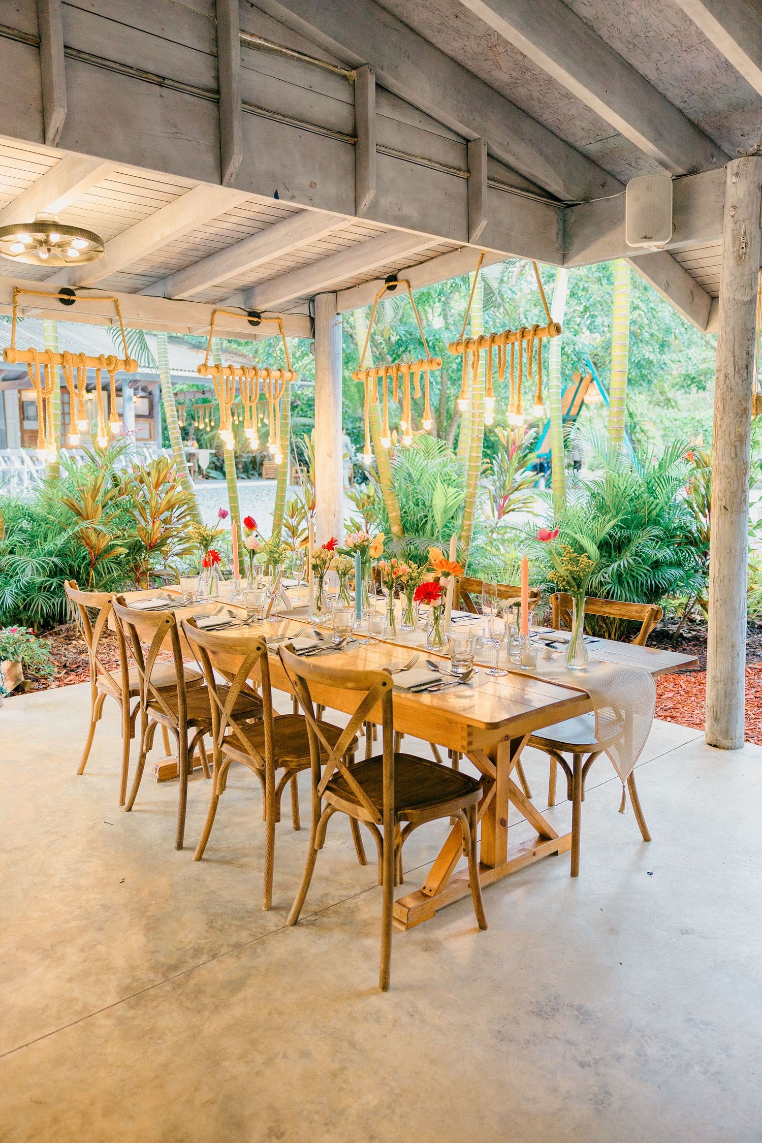 Rustic outdoor wedding reception area with wooden crossback chairs and tropical greenery.
