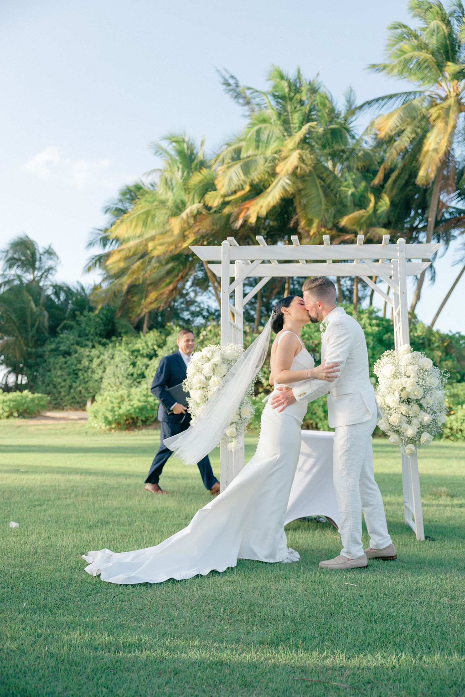Wedding ceremony on a lush green lawn with swaying palm trees and white floral decorations.