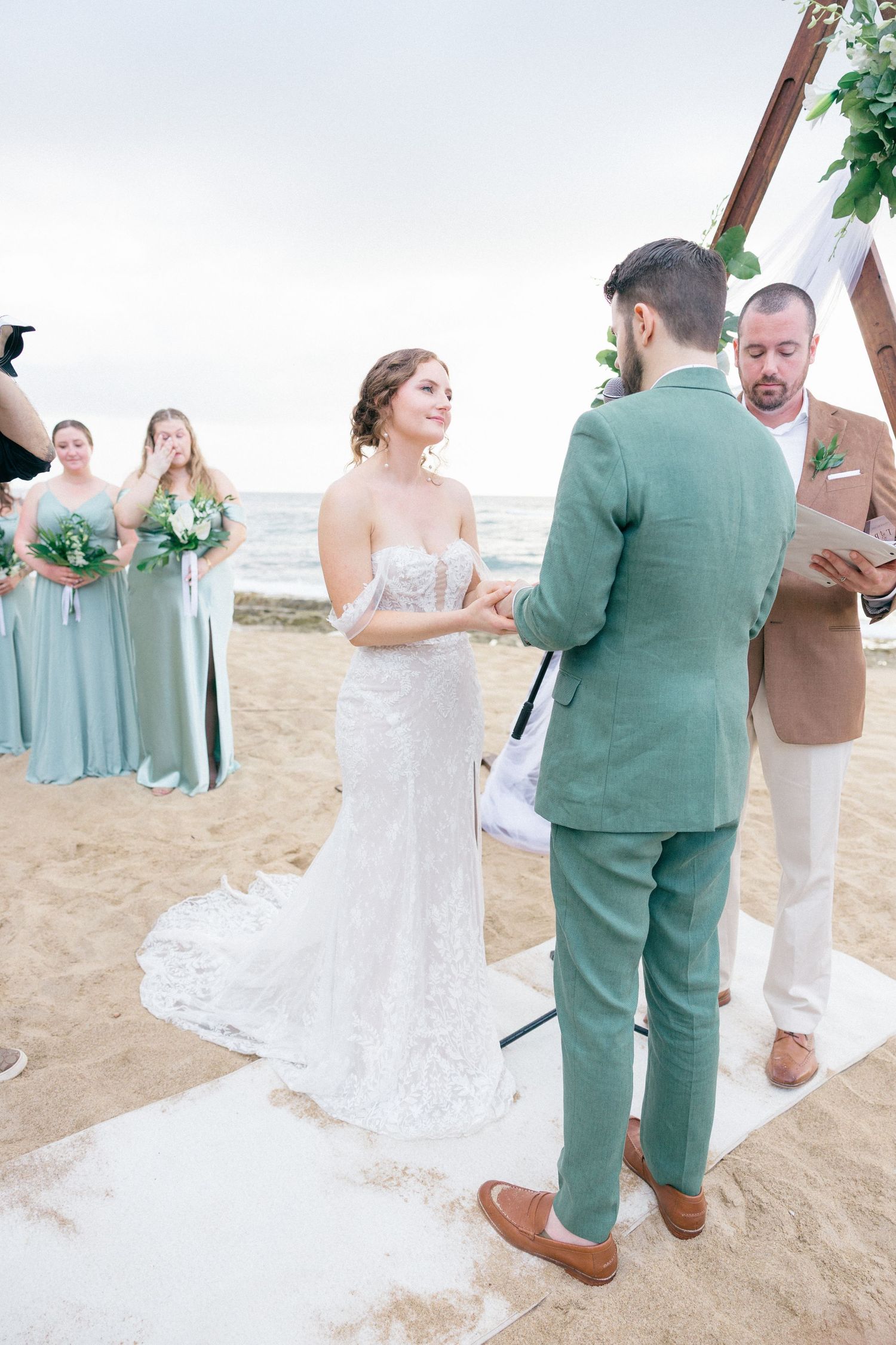 Beach wedding ceremony with bridesmaids in sage dresses and officiant in tan suit.