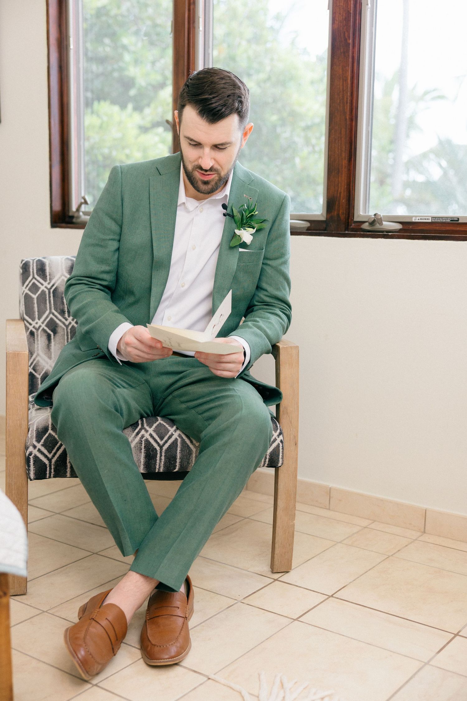 Groom in green suit reading letter while seated in modern chair by window.