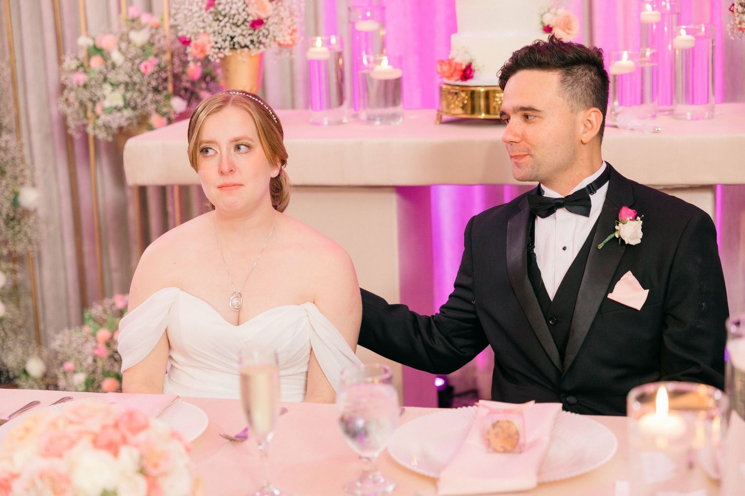Couple seated at decorated sweetheart table with pink floral arrangements and soft lighting.