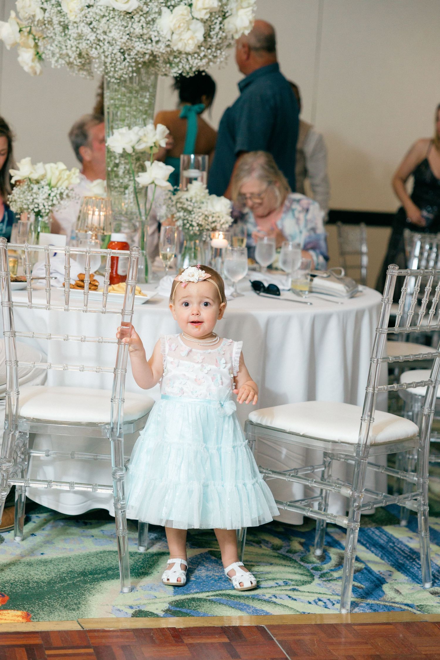 Little girl in light blue dress exploring wedding reception venue with clear chairs and white decor.