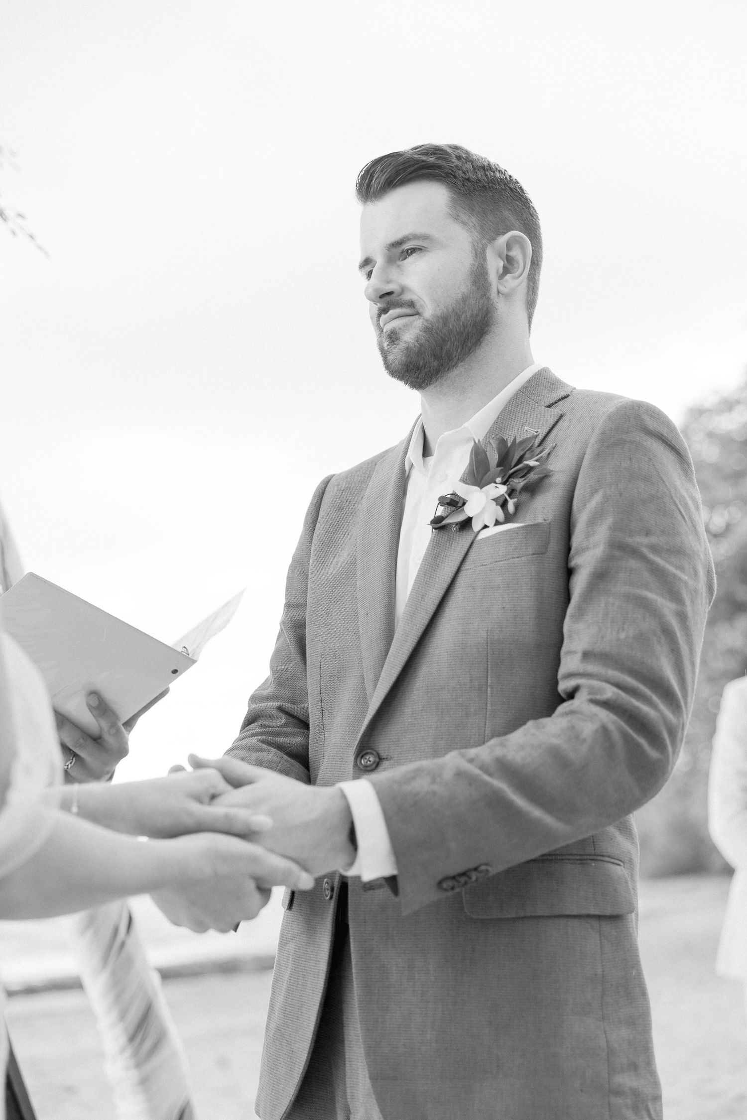 Groom in grey suit reads vows from paper during outdoor ceremony.