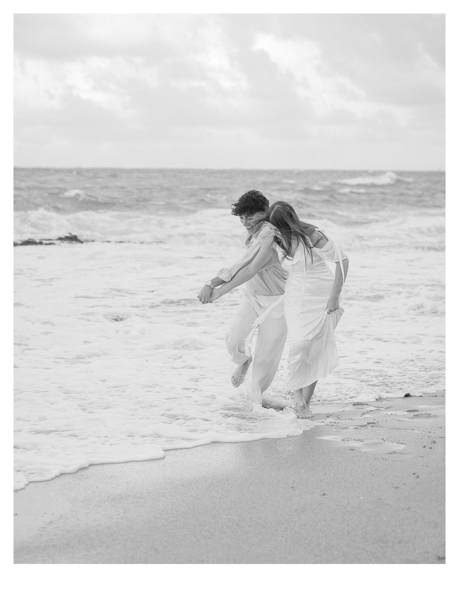 Black and white photo of couple playing in ocean waves on sandy beach.