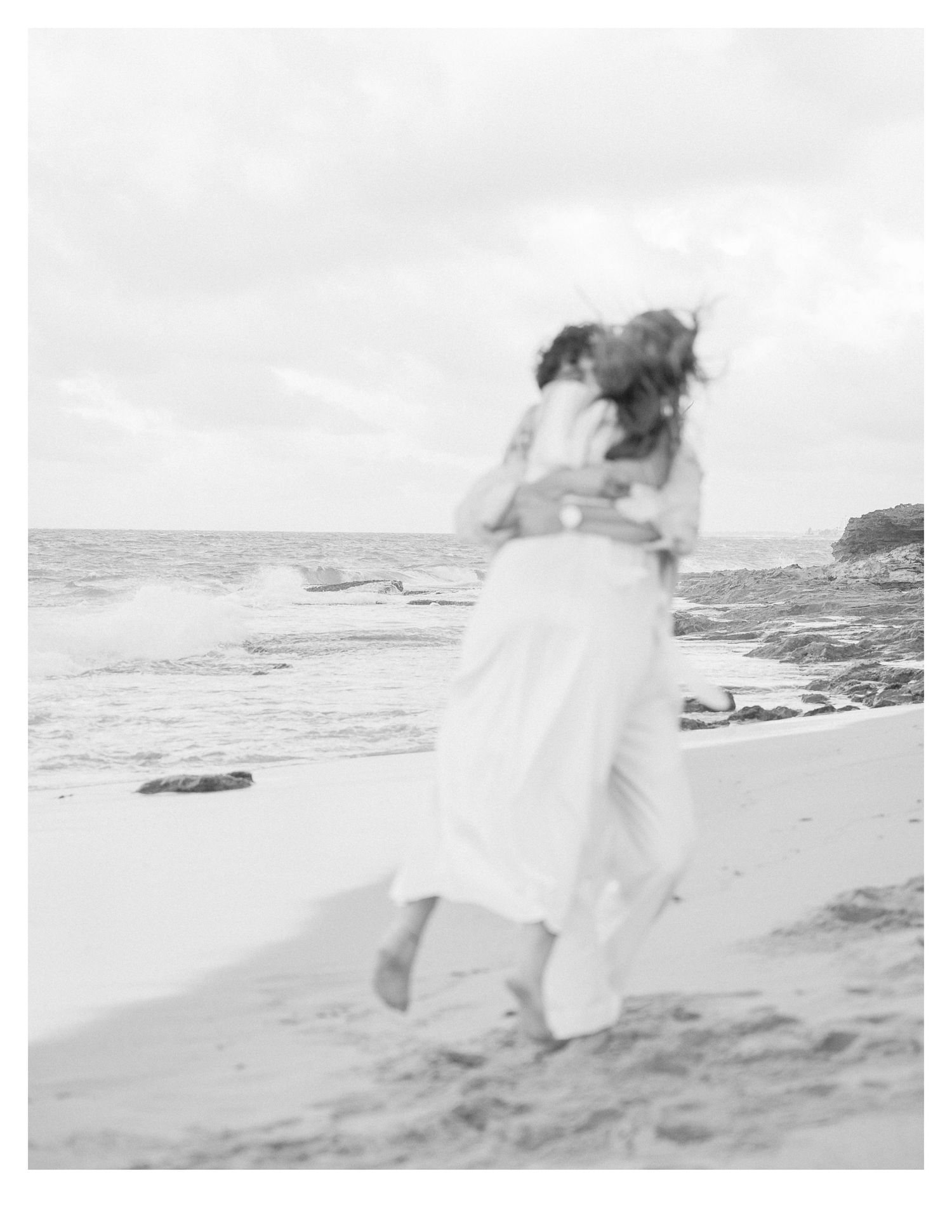 Black and white romantic beach photo of couple embracing by ocean waves.