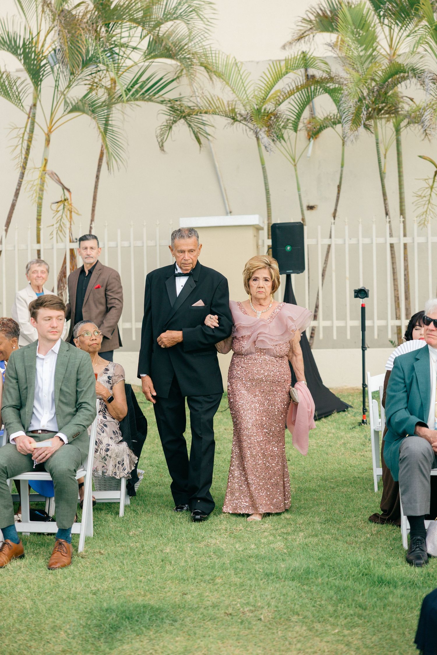 Wedding ceremony processional with guests seated in white chairs under tropical palm trees.