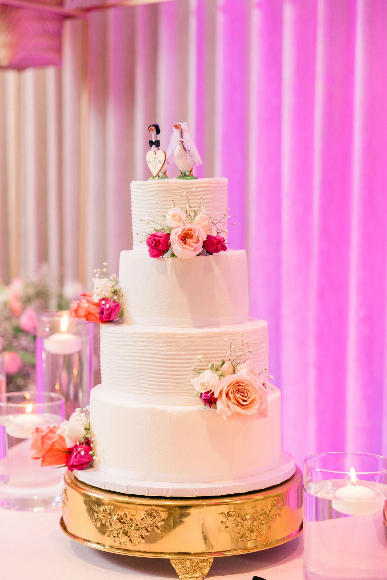 White tiered wedding cake with pink floral accents displayed against pink uplighting.