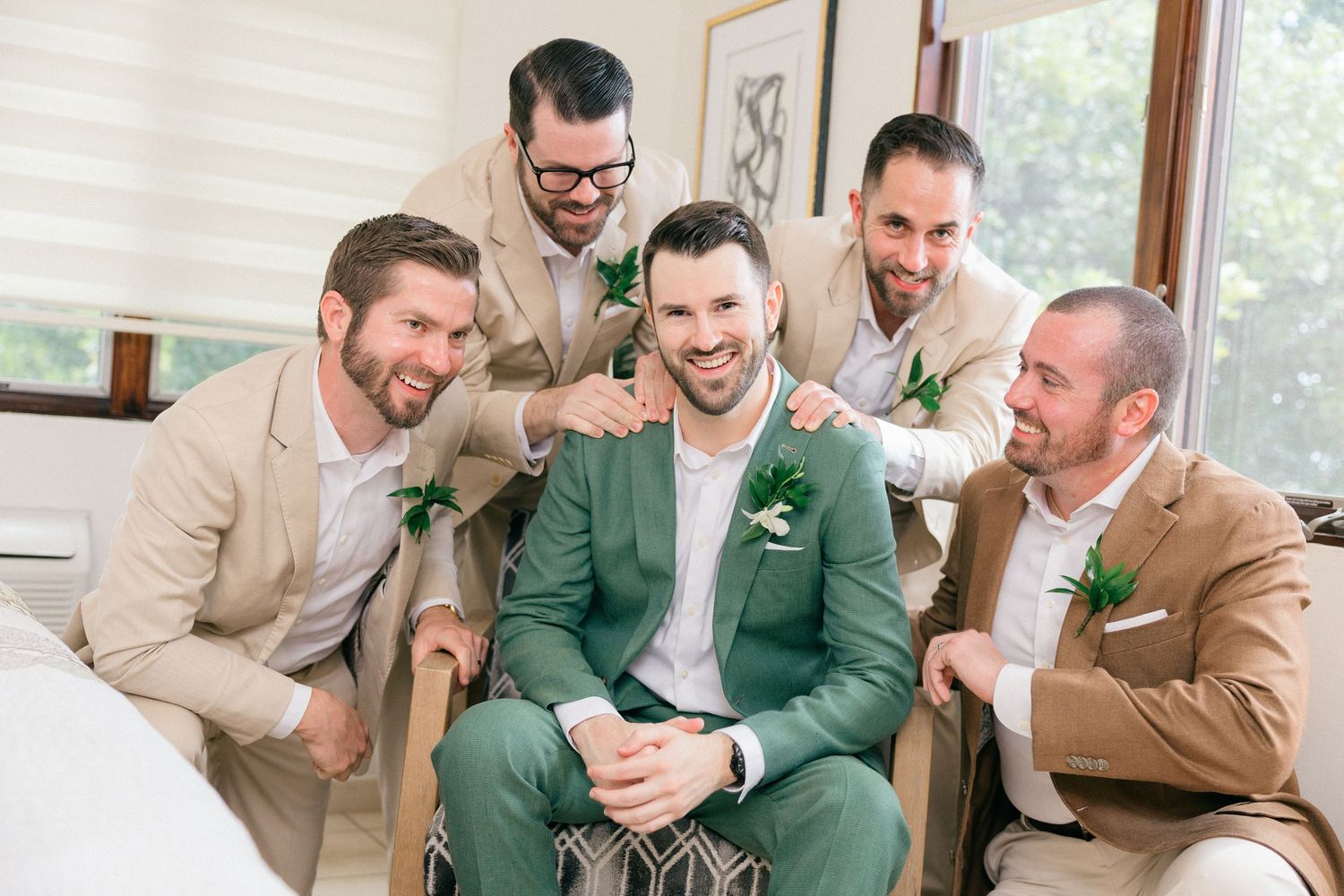Groomsmen in beige and green suits sharing laughs and celebrating together in bright indoor setting.