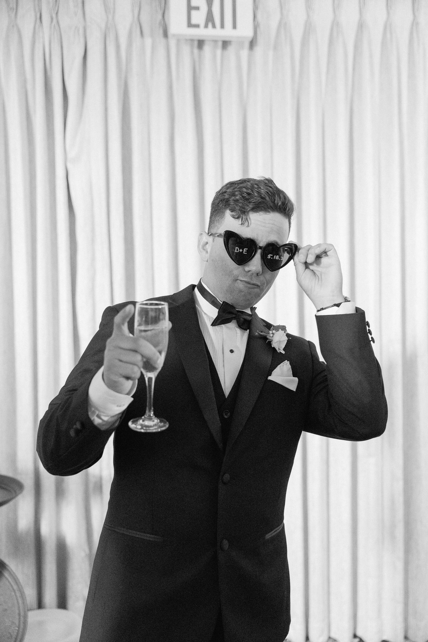 Man in tuxedo and sunglasses holding champagne glass against white curtain backdrop.