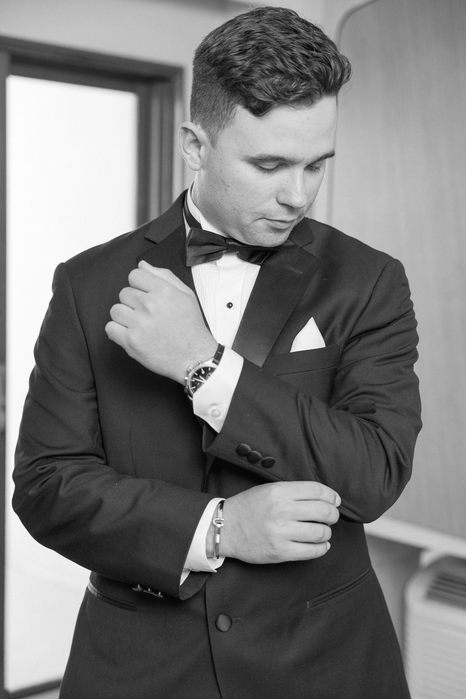 Groom adjusts his watch and cufflinks while wearing a classic black tuxedo.