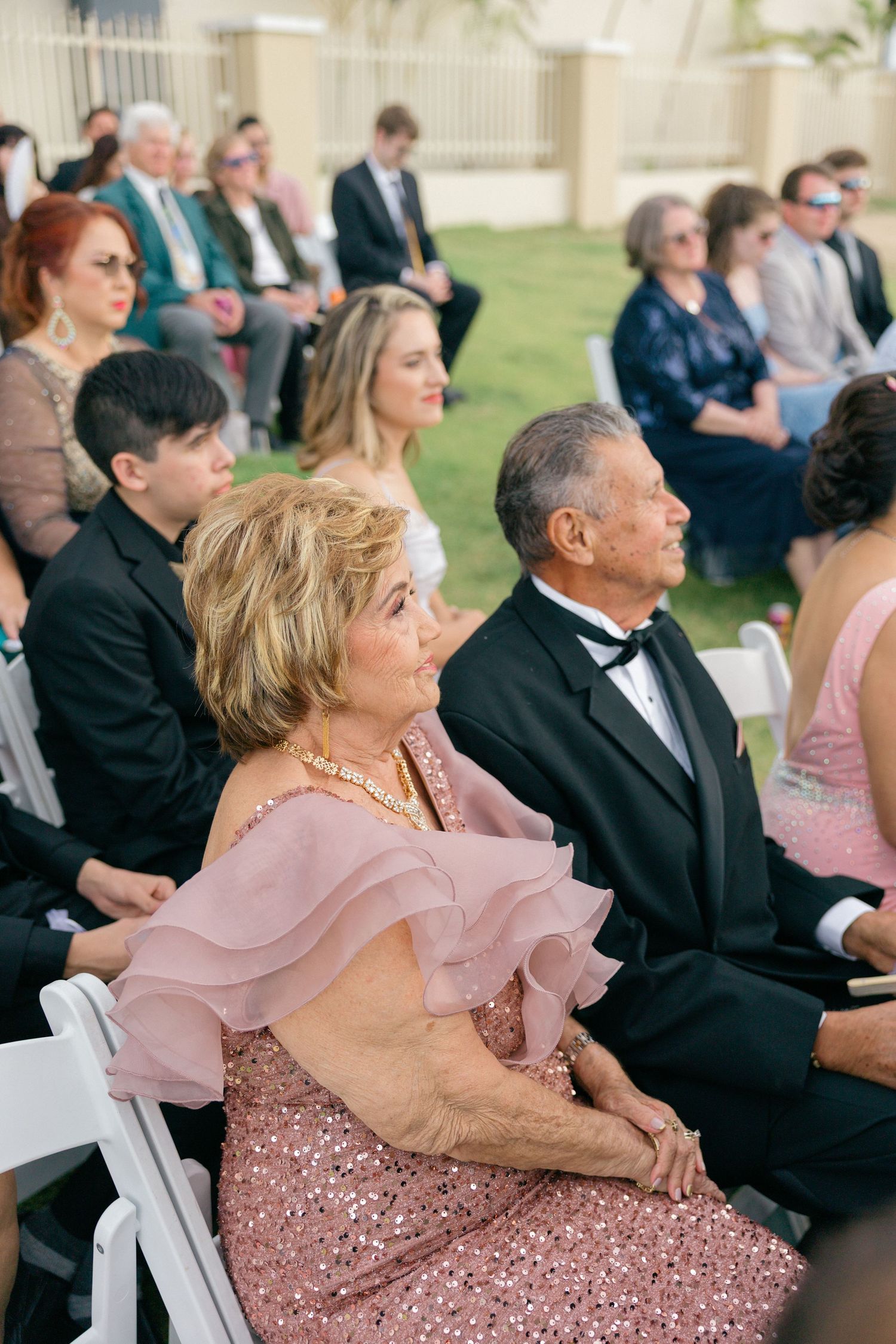 Wedding guests in formal attire seated on white chairs during outdoor ceremony.