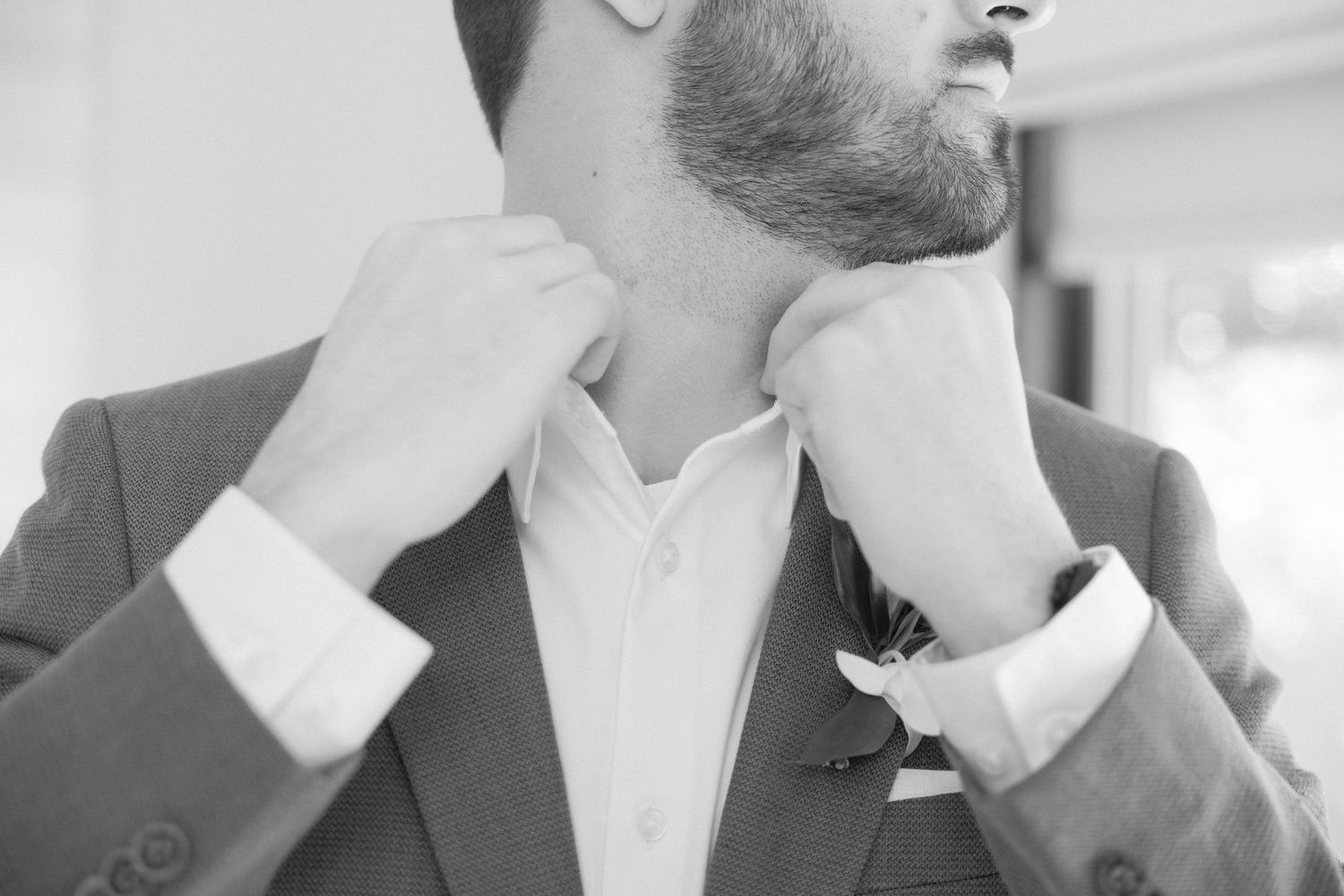 Black and white close-up of groom adjusting collar of formal attire.