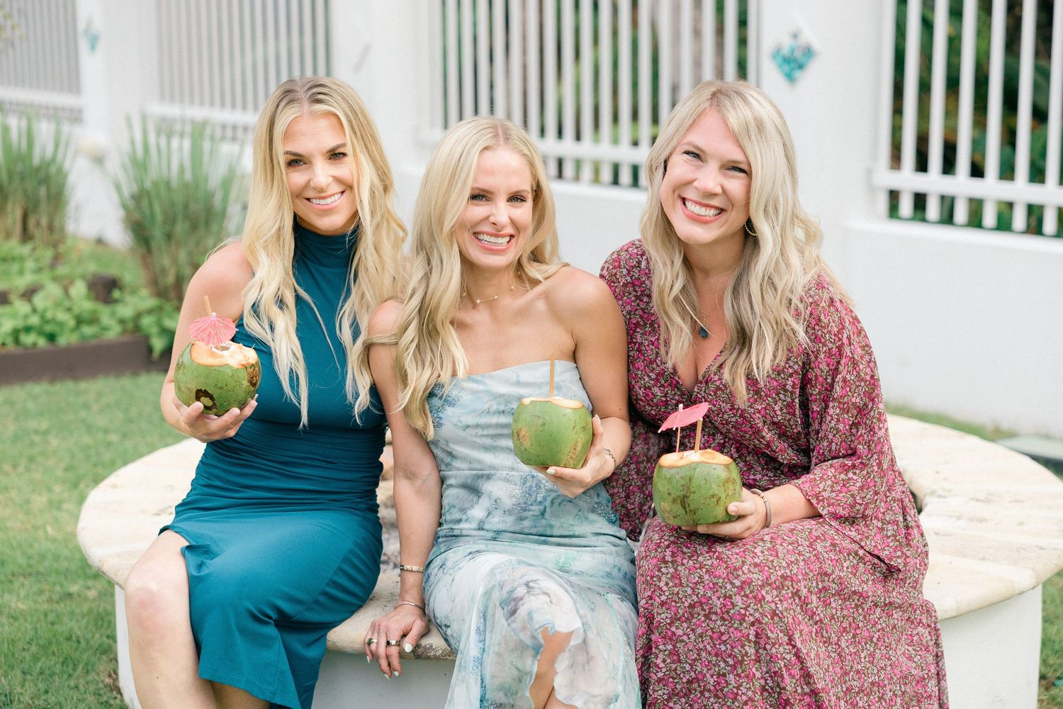 Three friends in elegant dresses sitting together holding coconut drinks.