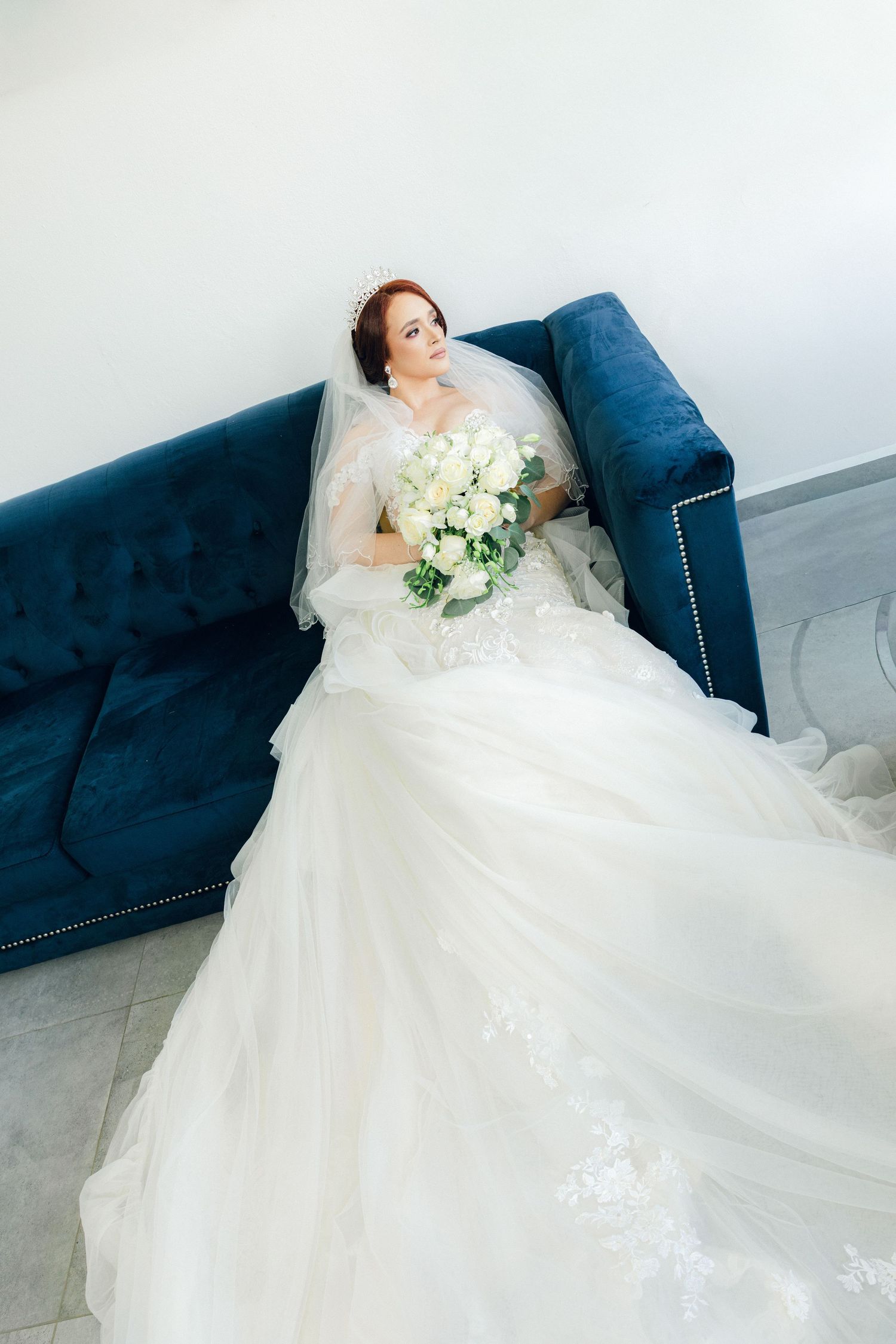 Bride in elegant white gown rests on blue velvet couch with floral bouquet.