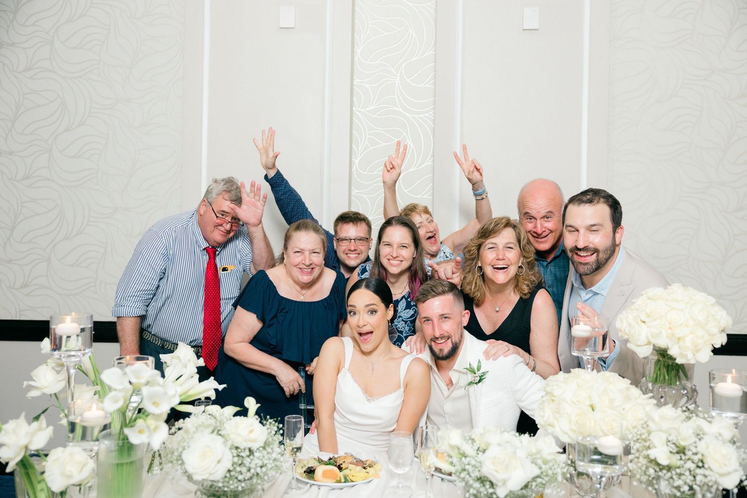 A festive group celebrates at an elegant wedding reception table decorated with white floral arrangements.