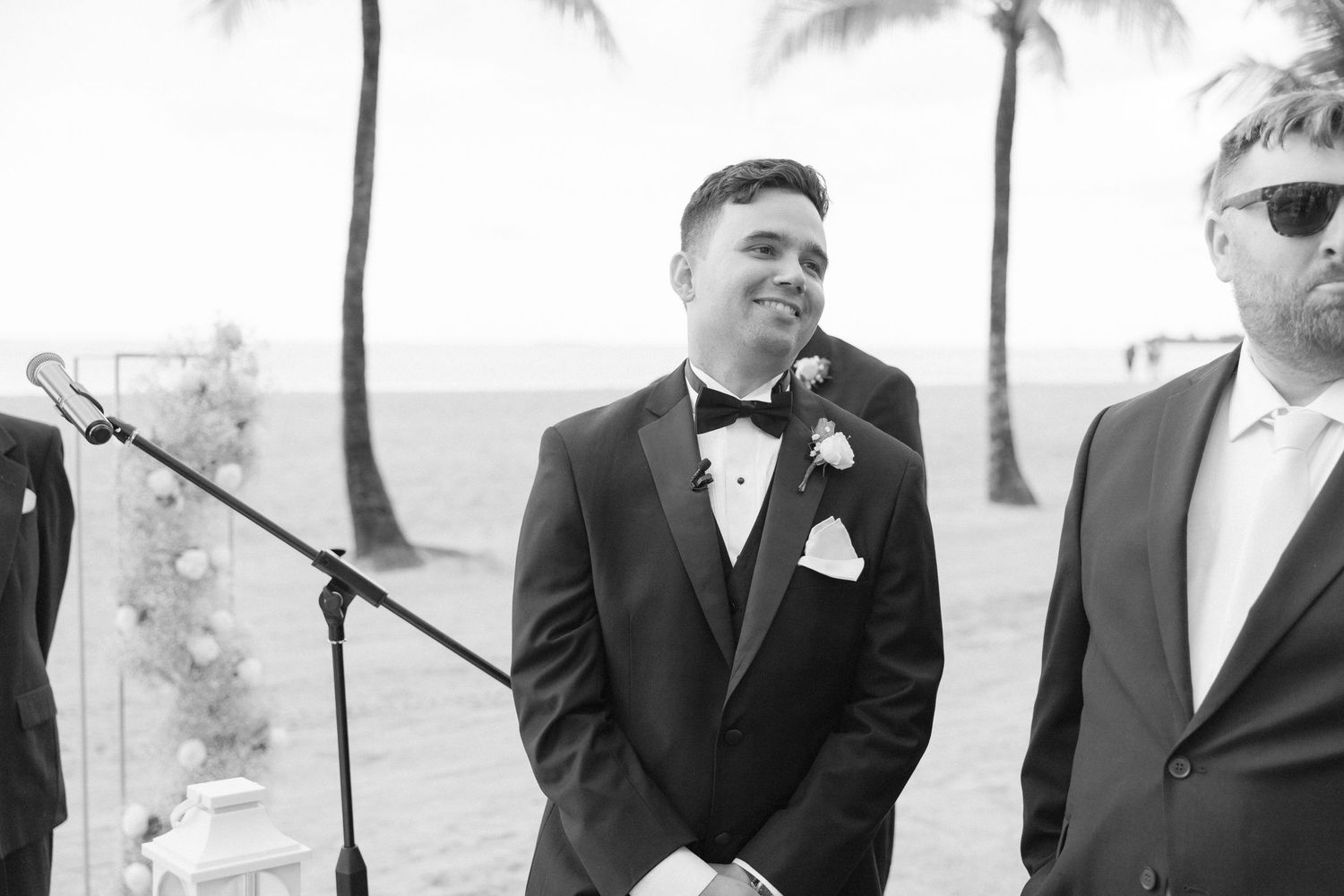 Wedding party members in formal attire smile and laugh together on a sunny beach with palm trees.