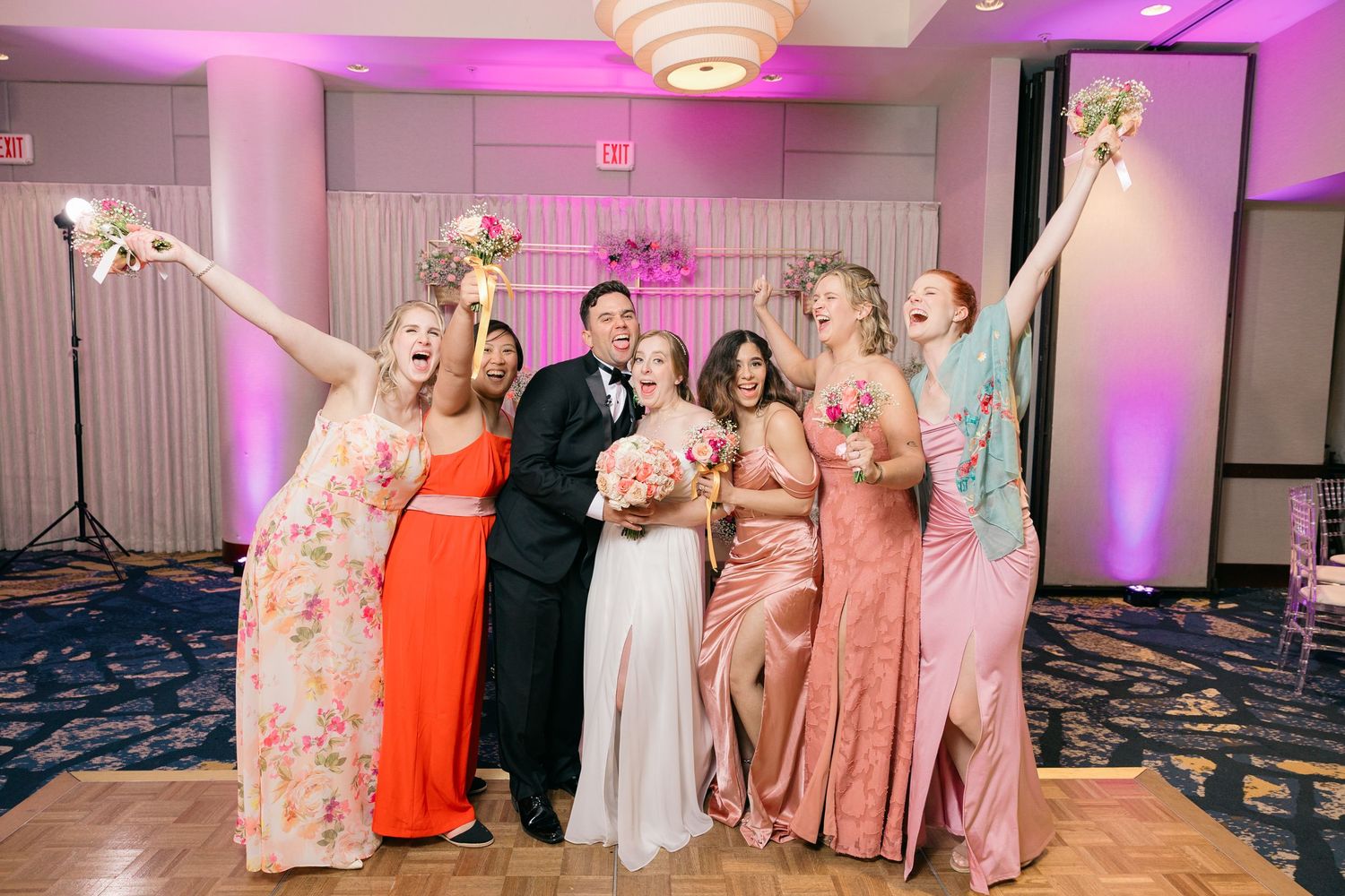 Wedding party poses energetically against pink-lit backdrop with floral decorations.