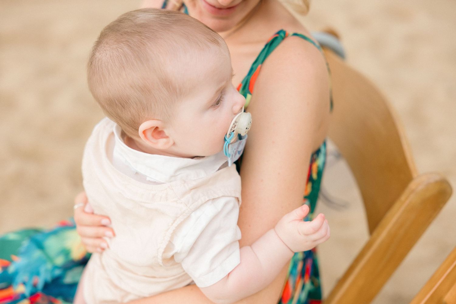 Baby in white dress sitting on wooden chairs at outdoor event.