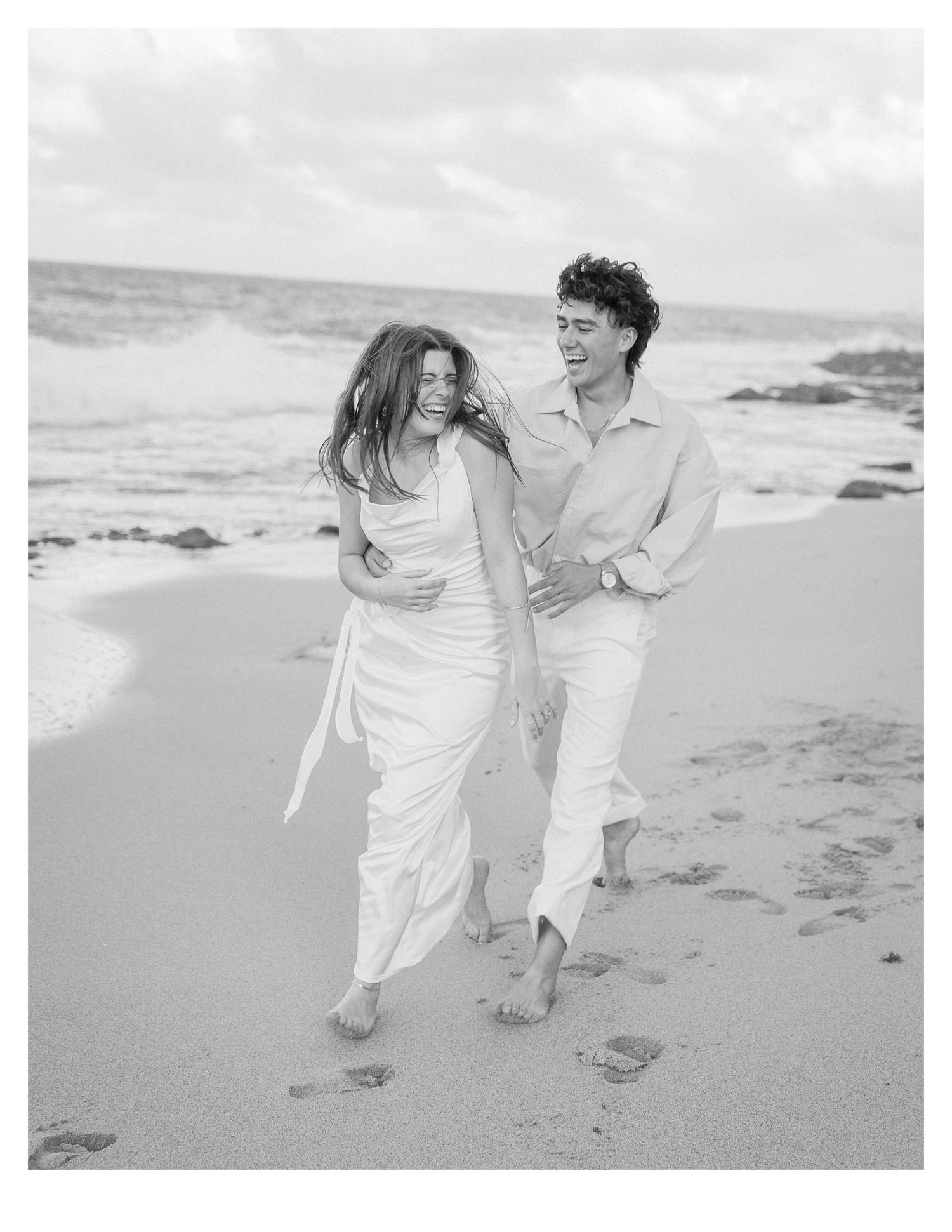 Candid beach photo of couple laughing while walking barefoot on sand.