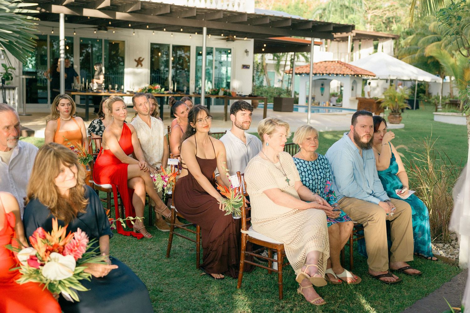 Wedding guests seated outdoors at long tables under trees for a garden reception celebration.