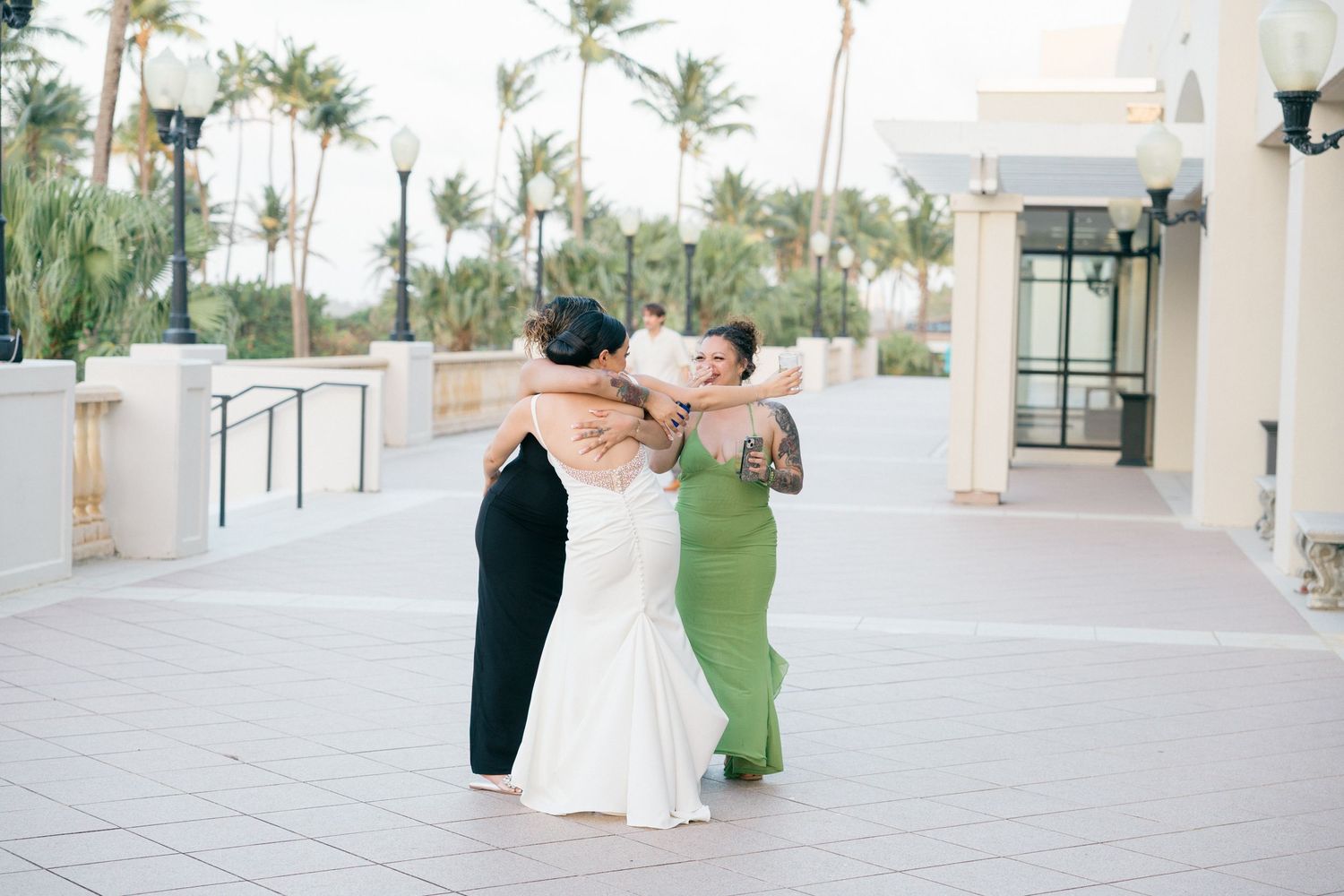Three people embrace in a group hug on an outdoor terrace with palm trees in the background.
