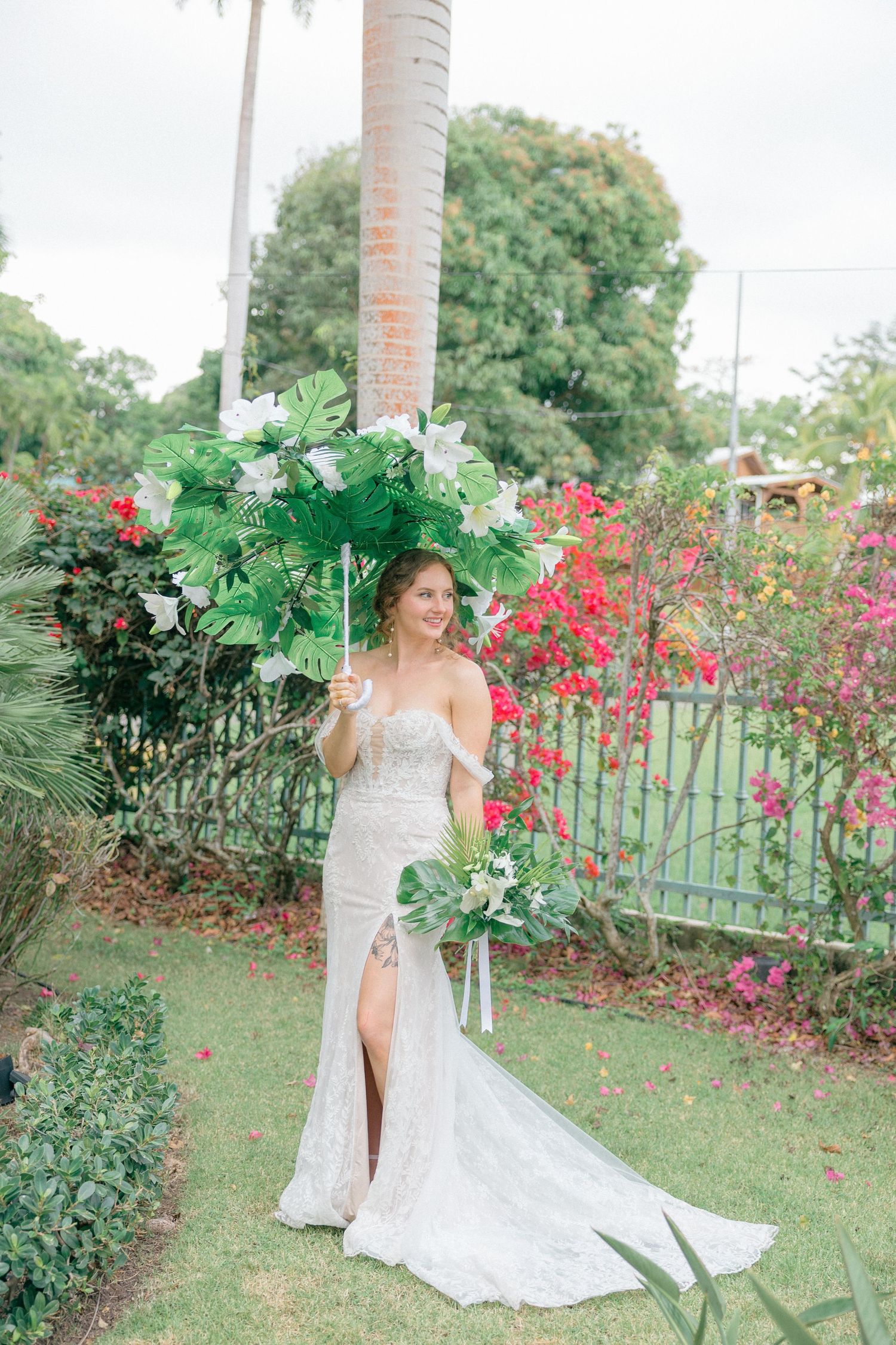 A white wedding dress flows elegantly in a tropical garden setting with vibrant red flowers and palm trees.