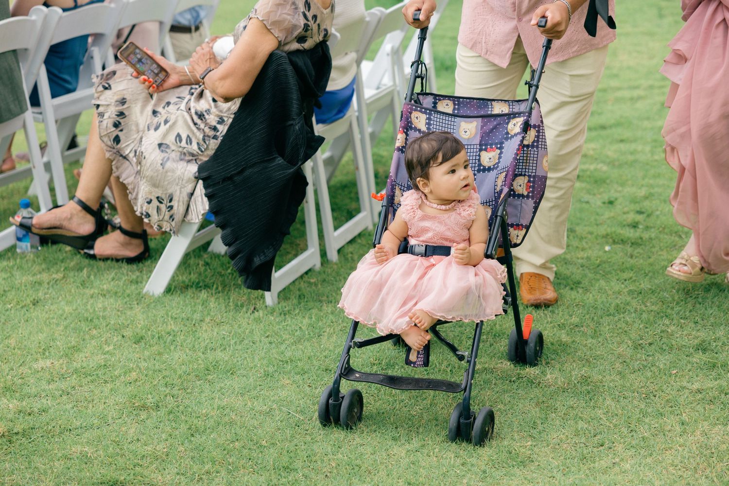 Small child in pink dress sits in stroller during outdoor wedding ceremony.