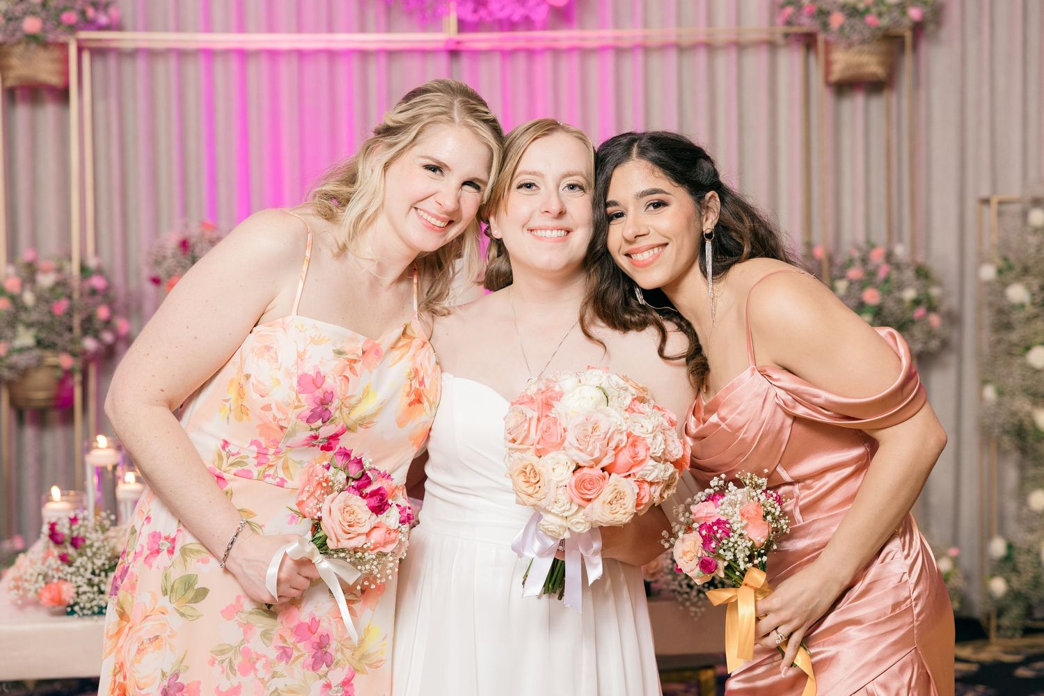 Three bridesmaids in floral and peach dresses pose with bouquets against a floral backdrop.