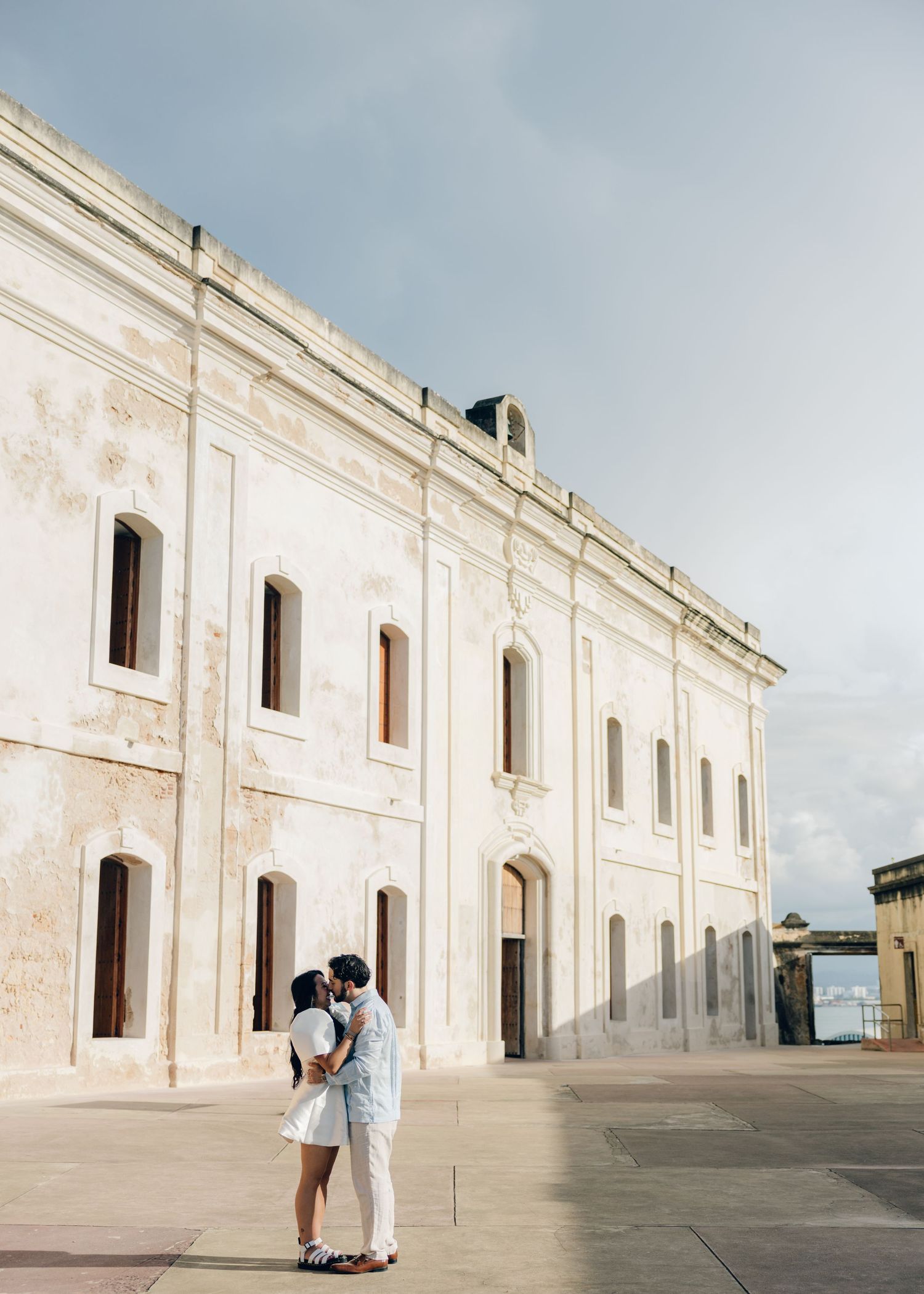 Couple kissing in a sunlit courtyard in front of a weathered historic stone building with arched windows under a soft cloudy sky.