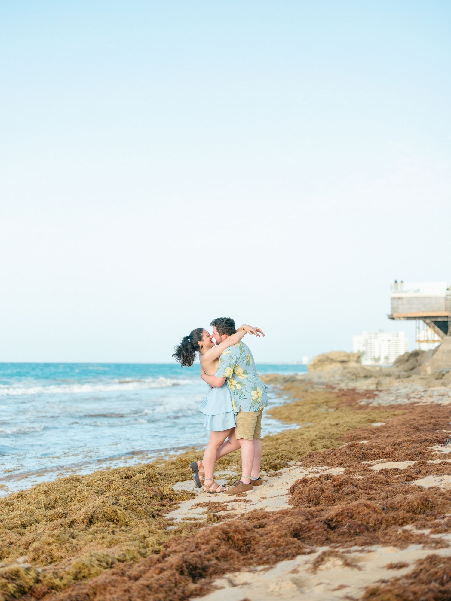 Couple sharing intimate moments on a beach with seaweed-covered shore.