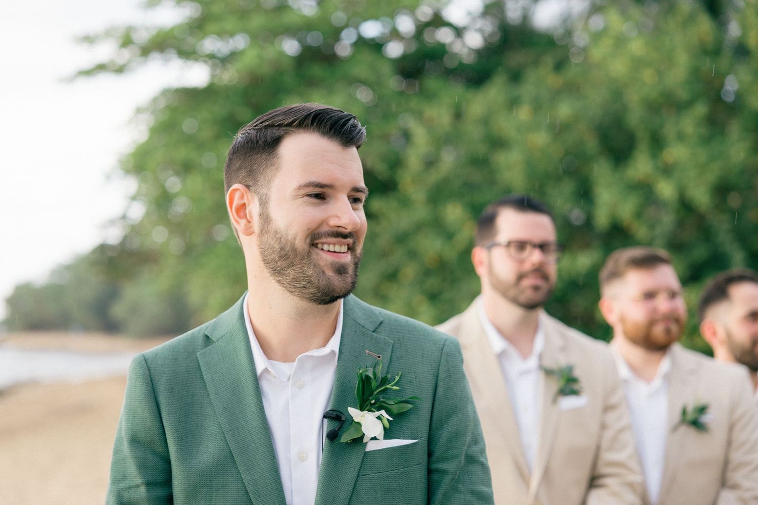 Wedding party members in formal attire pose on beach with green foliage background.