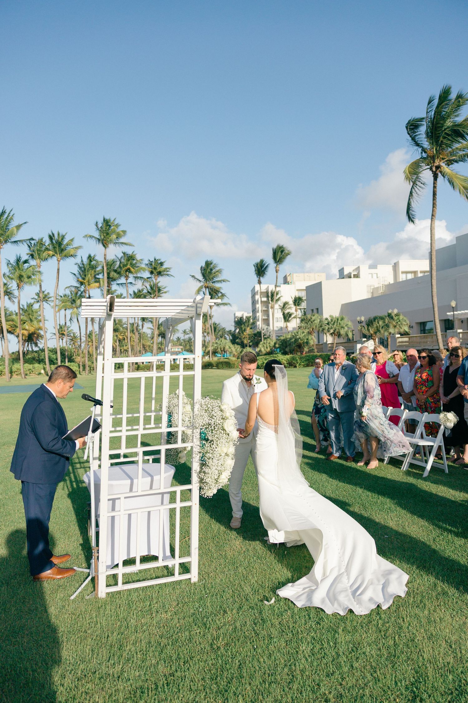 Wedding ceremony setup with white chairs and floral arrangements on a grassy lawn surrounded by palm trees.