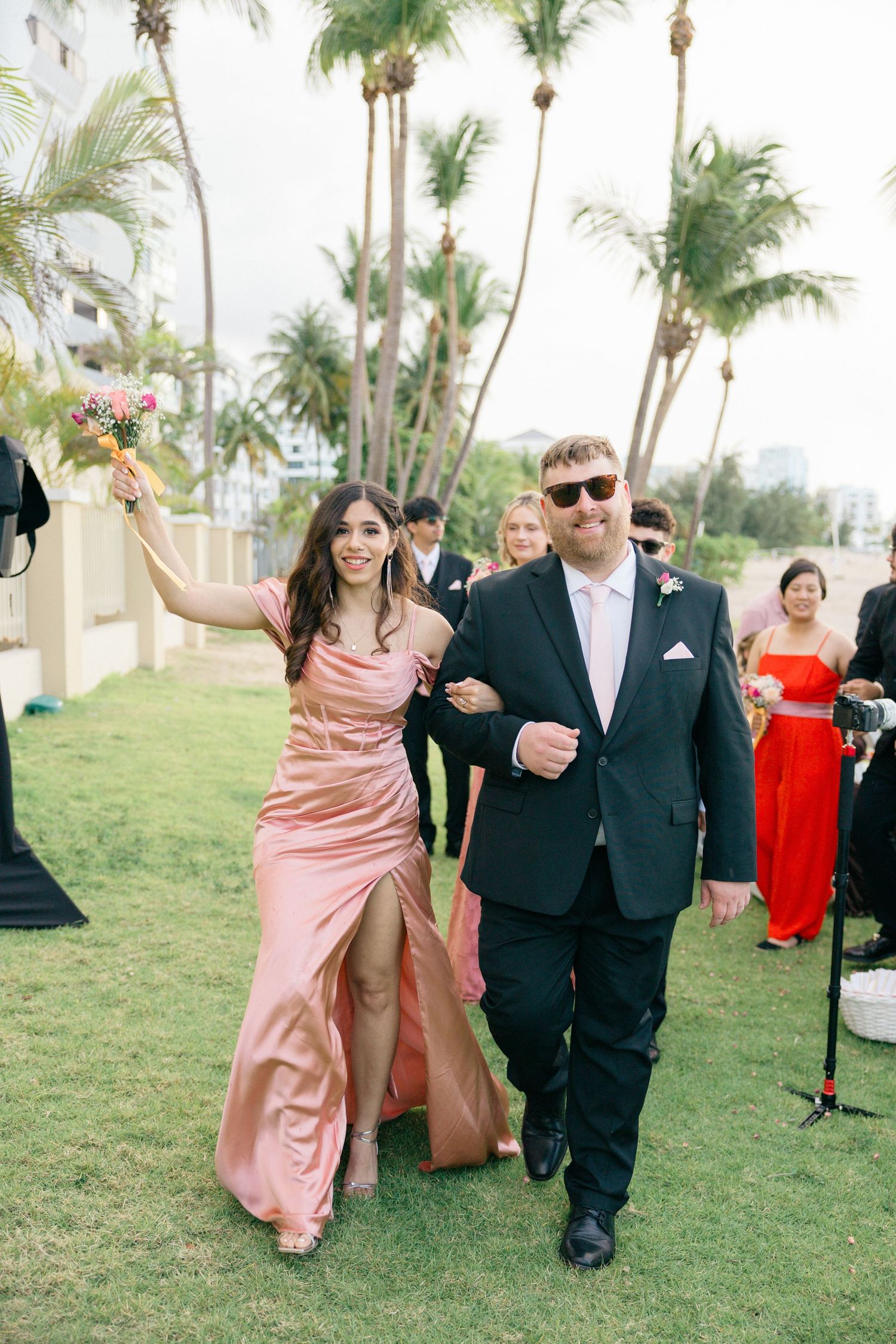 Wedding party members in formal attire walk across lawn during outdoor celebration.